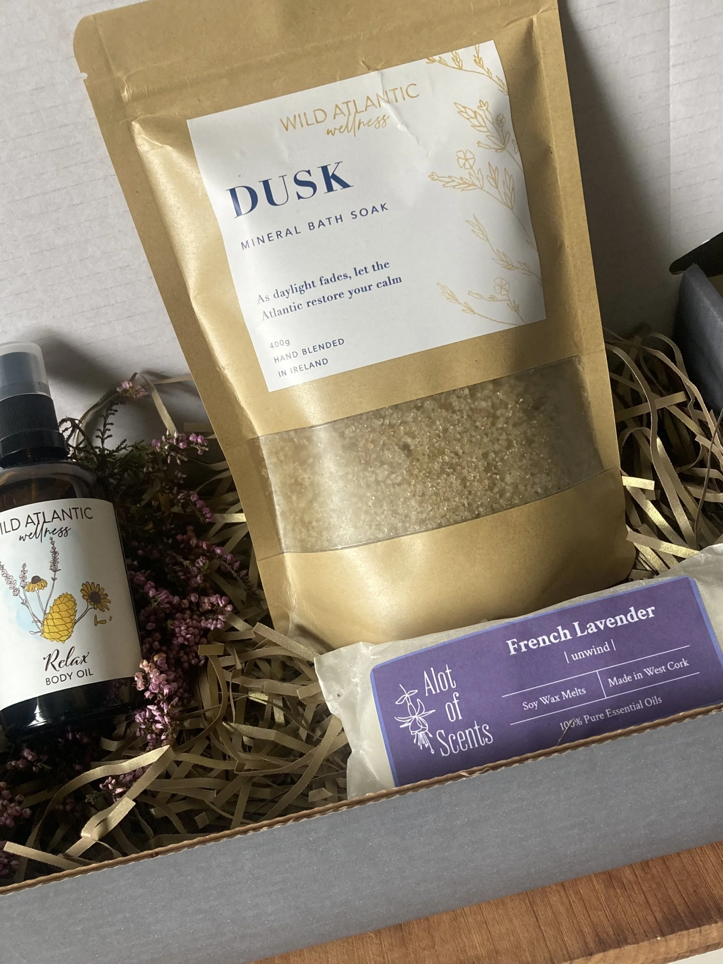 Angled view of the Rest & Restore gift set showing DUSK bath soak, Relax Body Oil, and lavender wax melt on a wooden board with dried heather