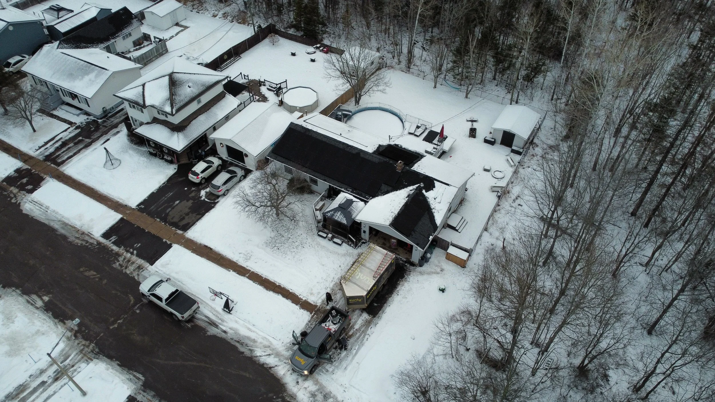 Aerial view of a snow-covered residential neighborhood with houses, cars, driveways, and trees.