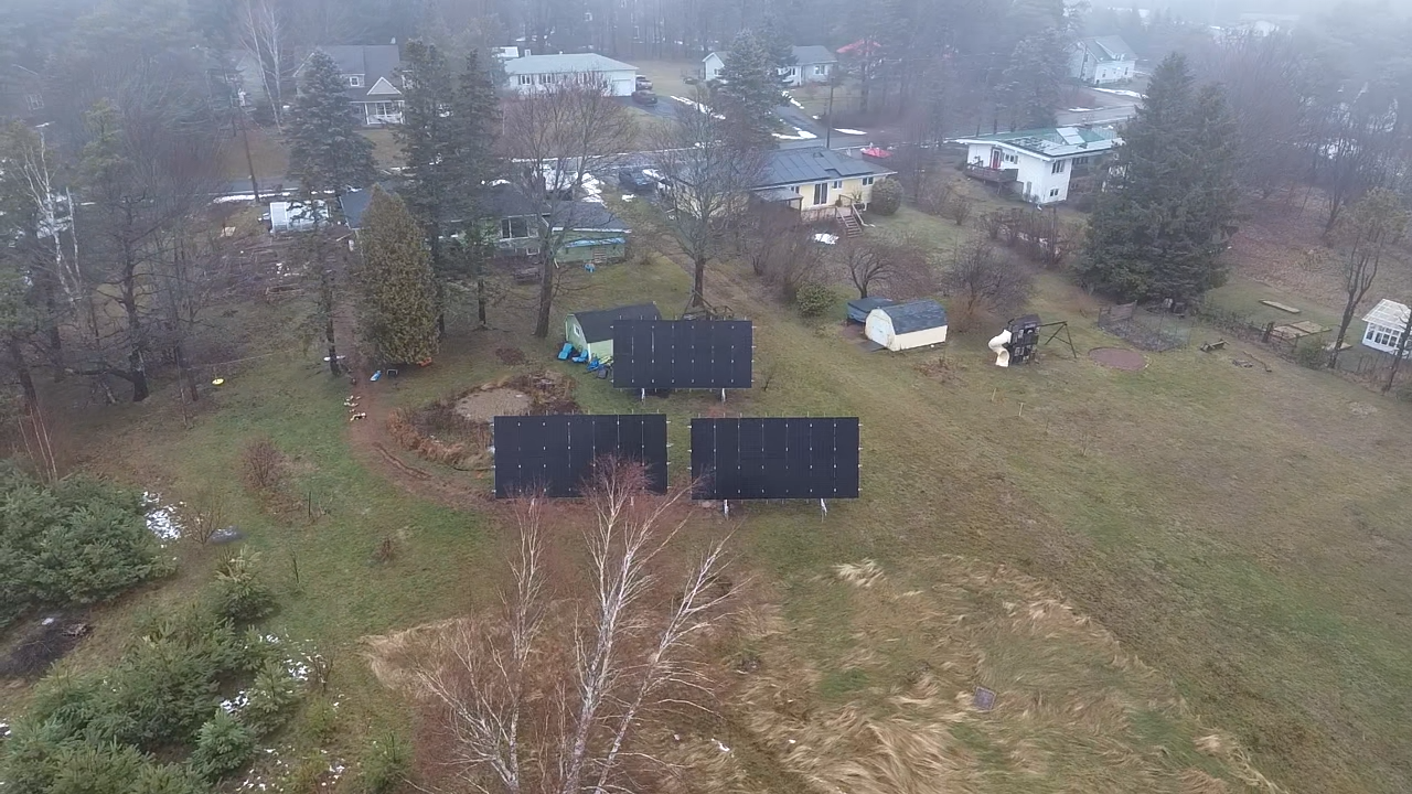 Aerial view of a suburban backyard with solar panels, trees, sheds, and neighboring houses.