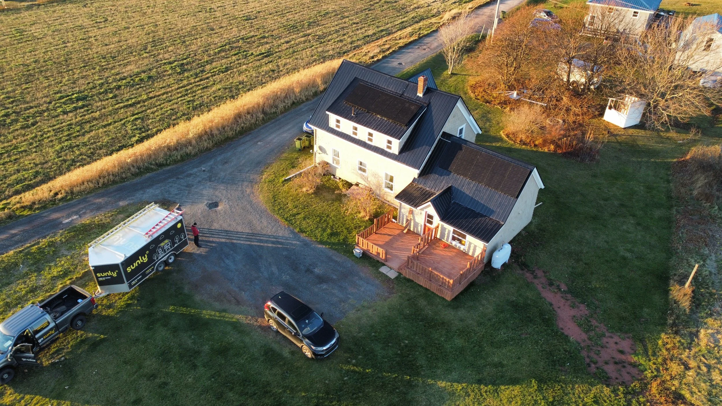 Aerial view of a two-story house with solar panels on the roof, a wooden deck at the back, and a yard surrounded by fields and trees. A truck with solar panel graphics and a person are parked nearby.