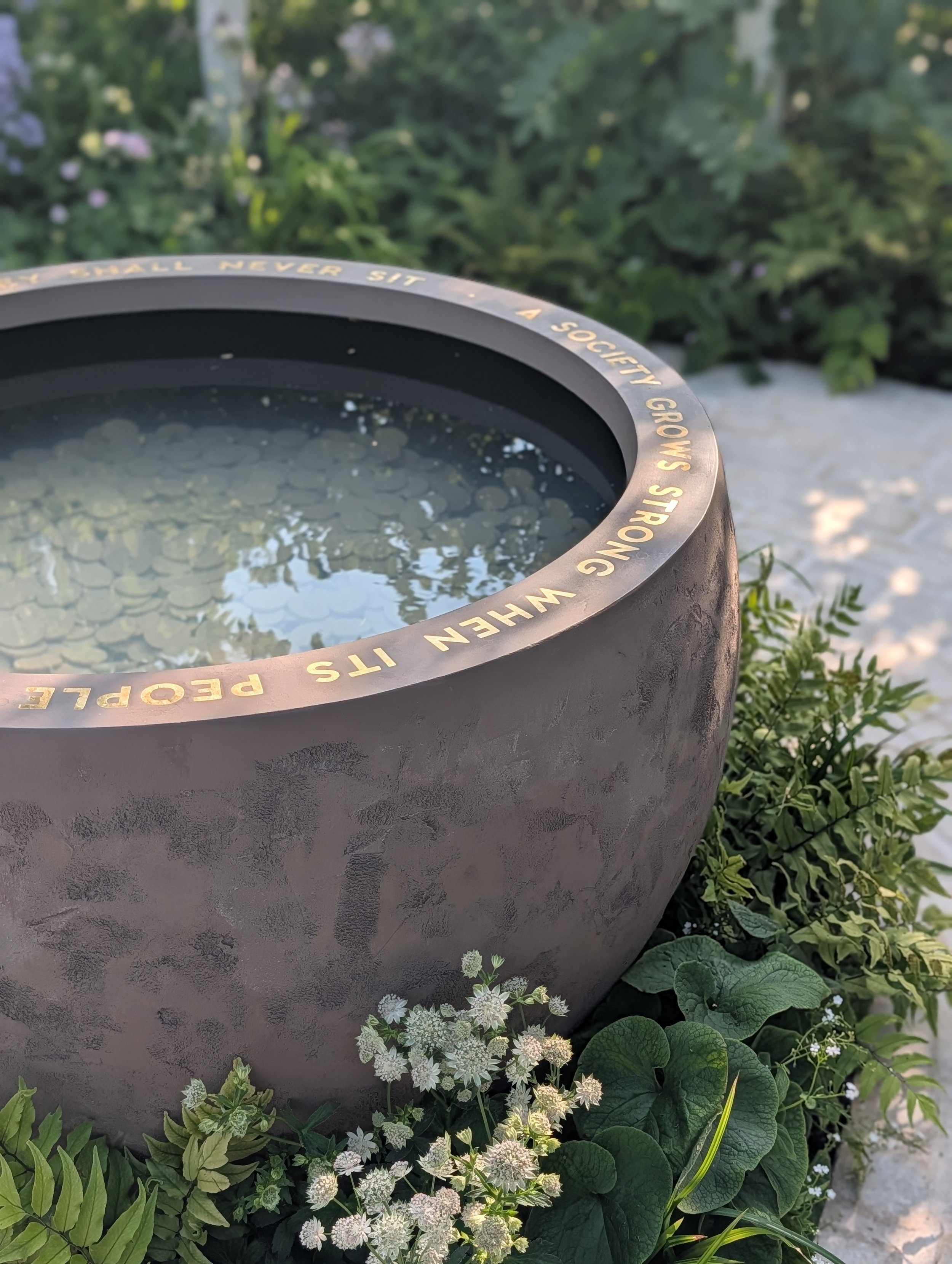 A round, concrete planter with an inscription around the rim, filled with water reflecting the sky, surrounded by green plants and small white flowers.