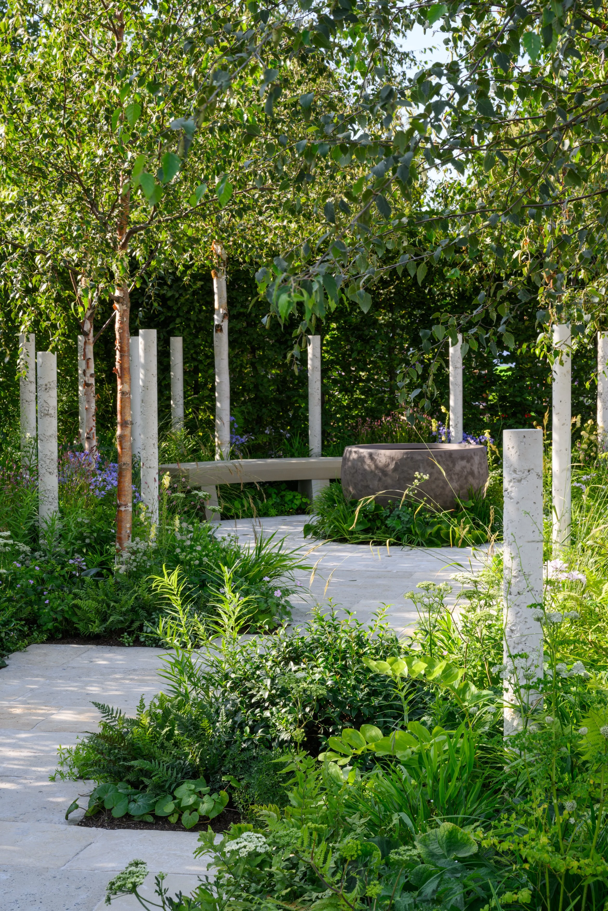 A lush garden with a stone pathway, green bushes, and small trees, surrounded by white-painted stakes and a black circular planter.