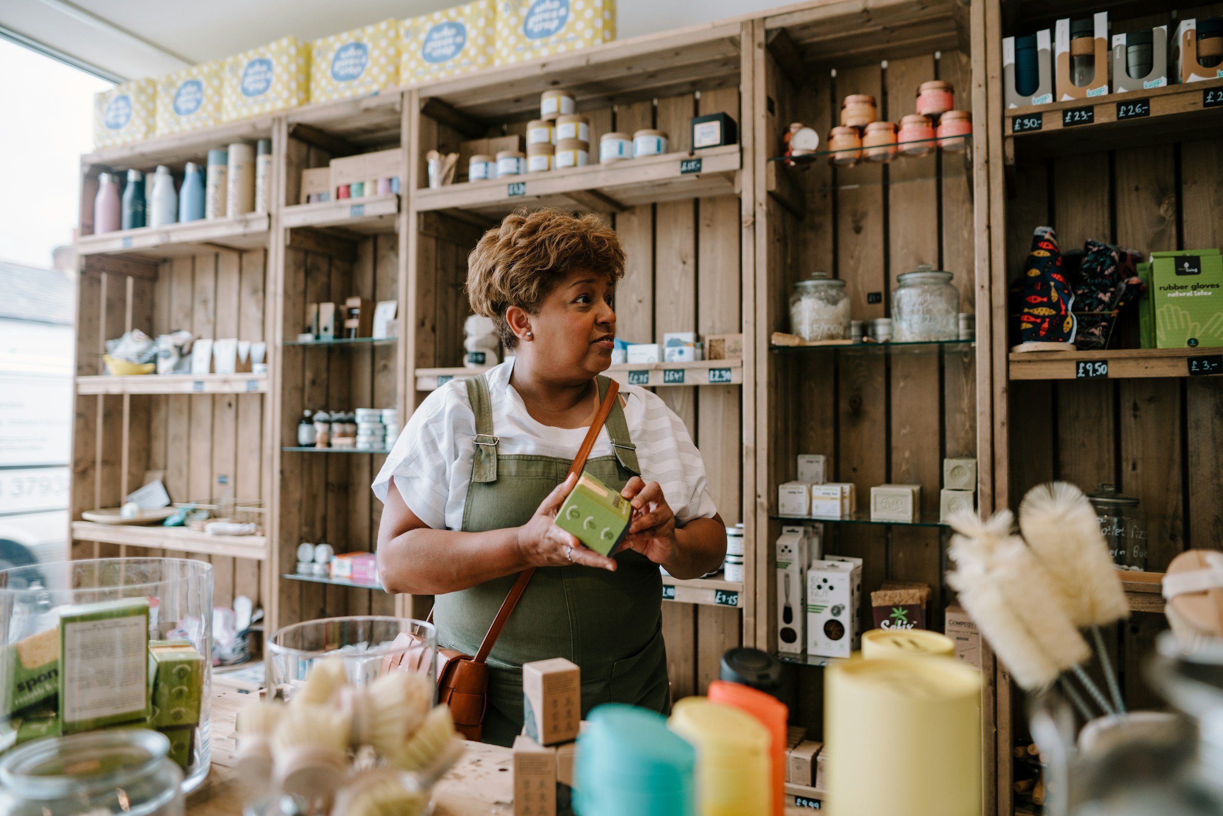 A person stands inside a small shop with wooden shelves, holding a green boxed product and looking to the side..jpg