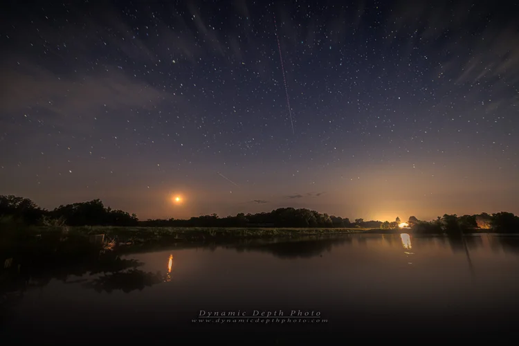 Stars and a bright moon shine above still water and its reflections.