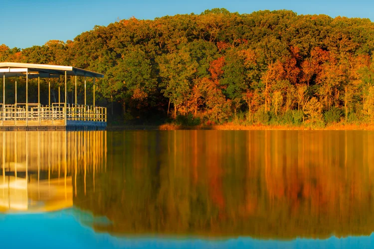 Orange and gold trees reflect beside a lakeside pavilion in still water.