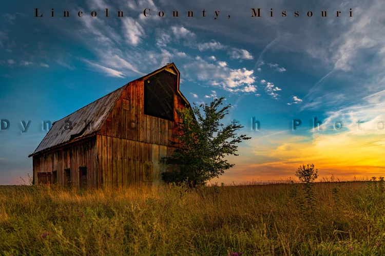 A weathered wooden barn stands against blue clouds and a warm sunset horizon.