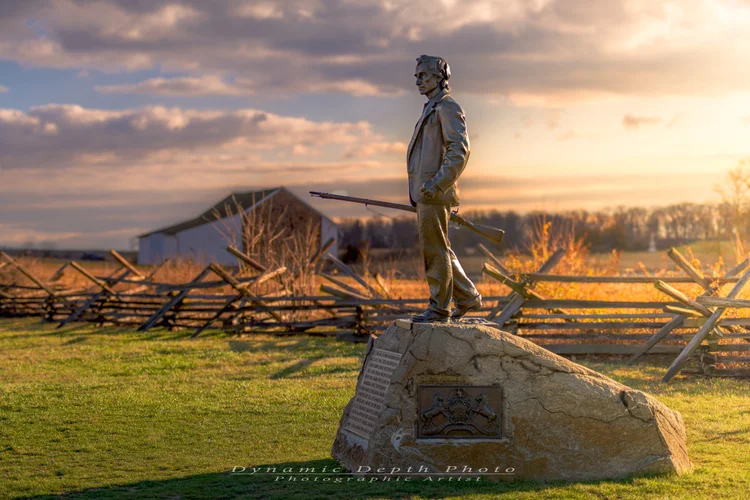 A soldier monument stands beside split-rail fencing and farm buildings in warm evening light.