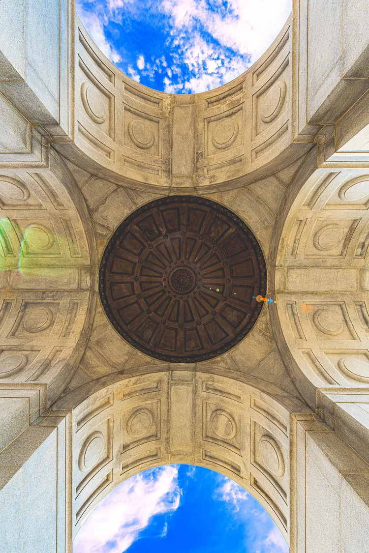 The interior arches and carved ceiling of a stone memorial frame patches of blue sky.