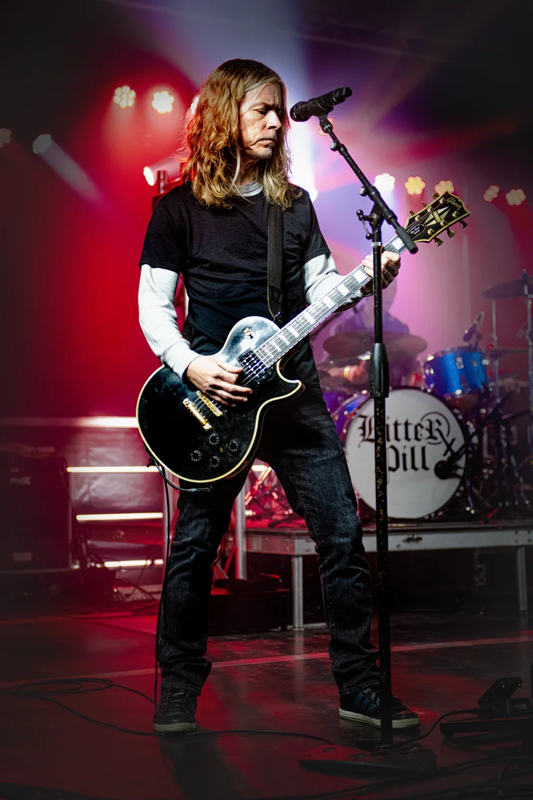 A guitarist performs at a microphone beneath red and purple stage lighting.