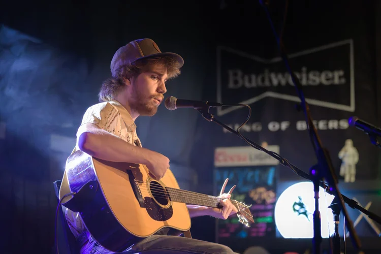 A musician in a cap performs with an acoustic guitar beneath blue stage lights.