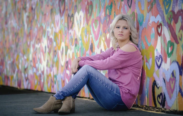 A woman in a pink sweater sits beside a brightly painted wall.