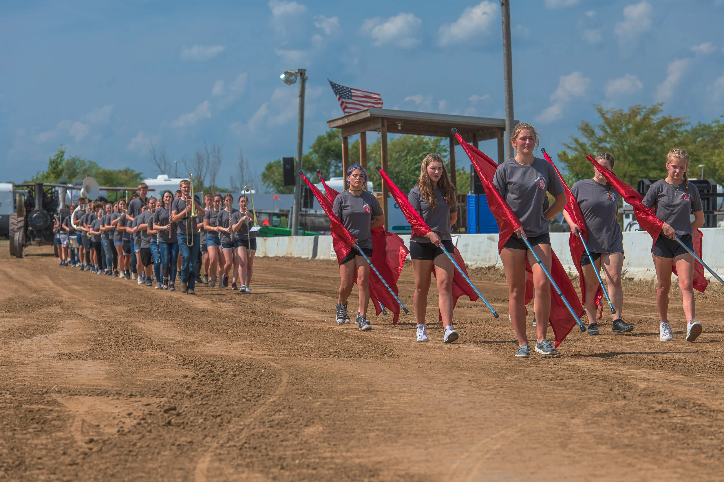 A flag team carrying red flags leads a marching band along the dirt track at the 2022 Old Thresher Days.