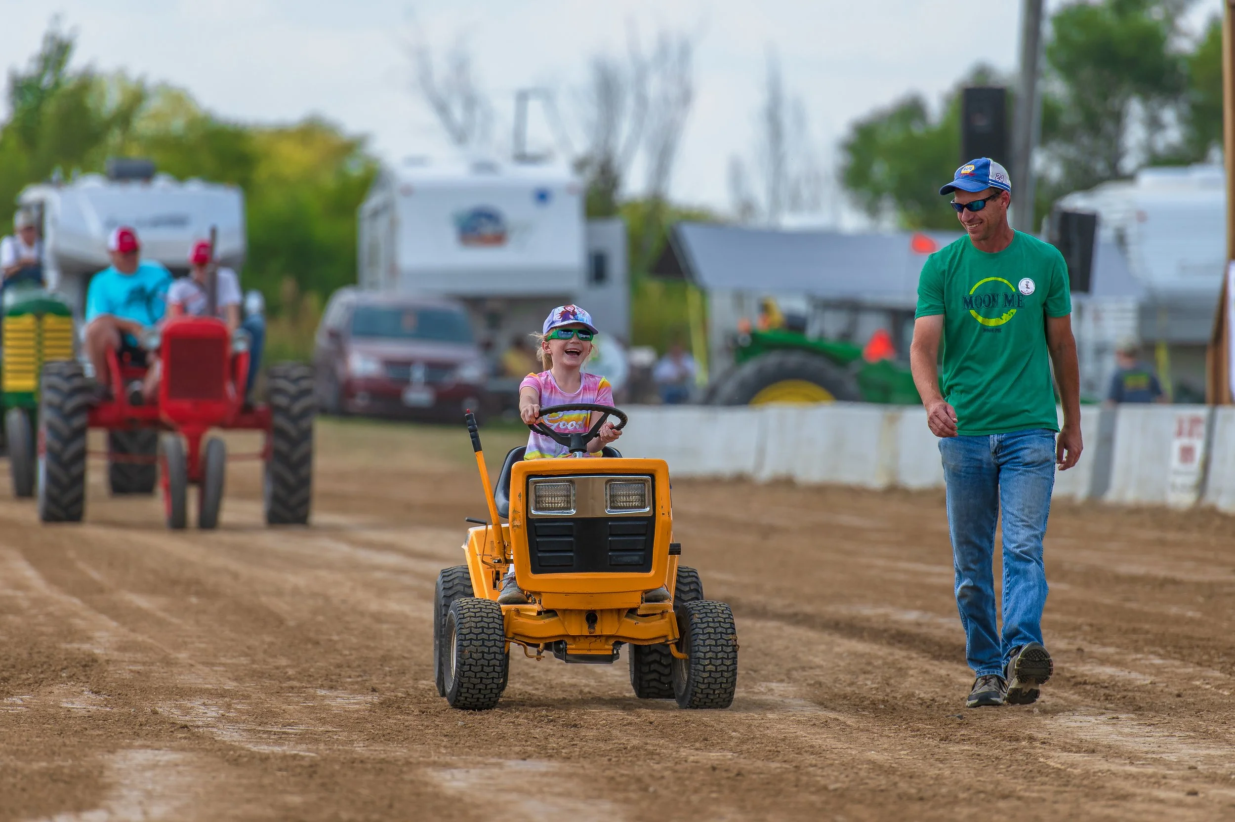 A small yellow lawn tractor travels beside a walking adult, with larger tractors behind, at the 2022 Old Thresher Days.