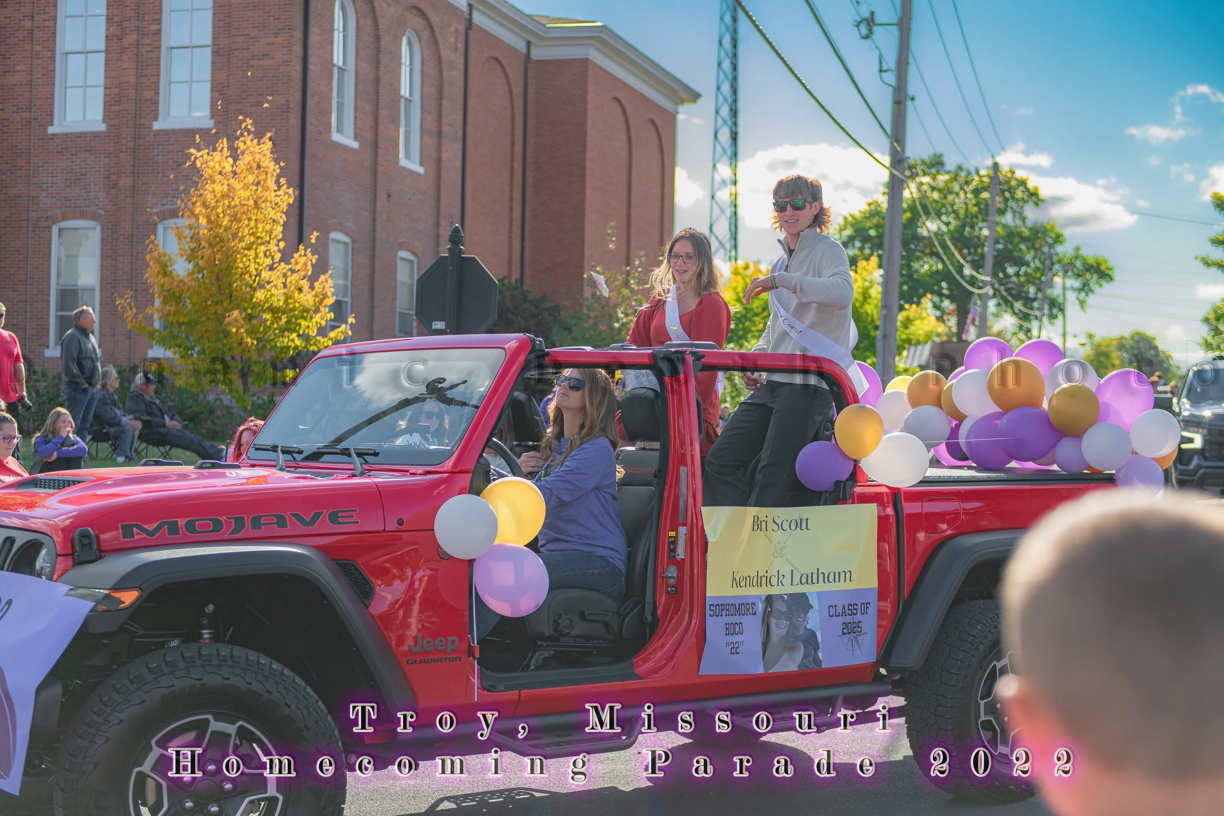 Two passengers wave from a red Jeep decorated with purple, gold, and white balloons during the 2022 Troy Homecoming Parade.