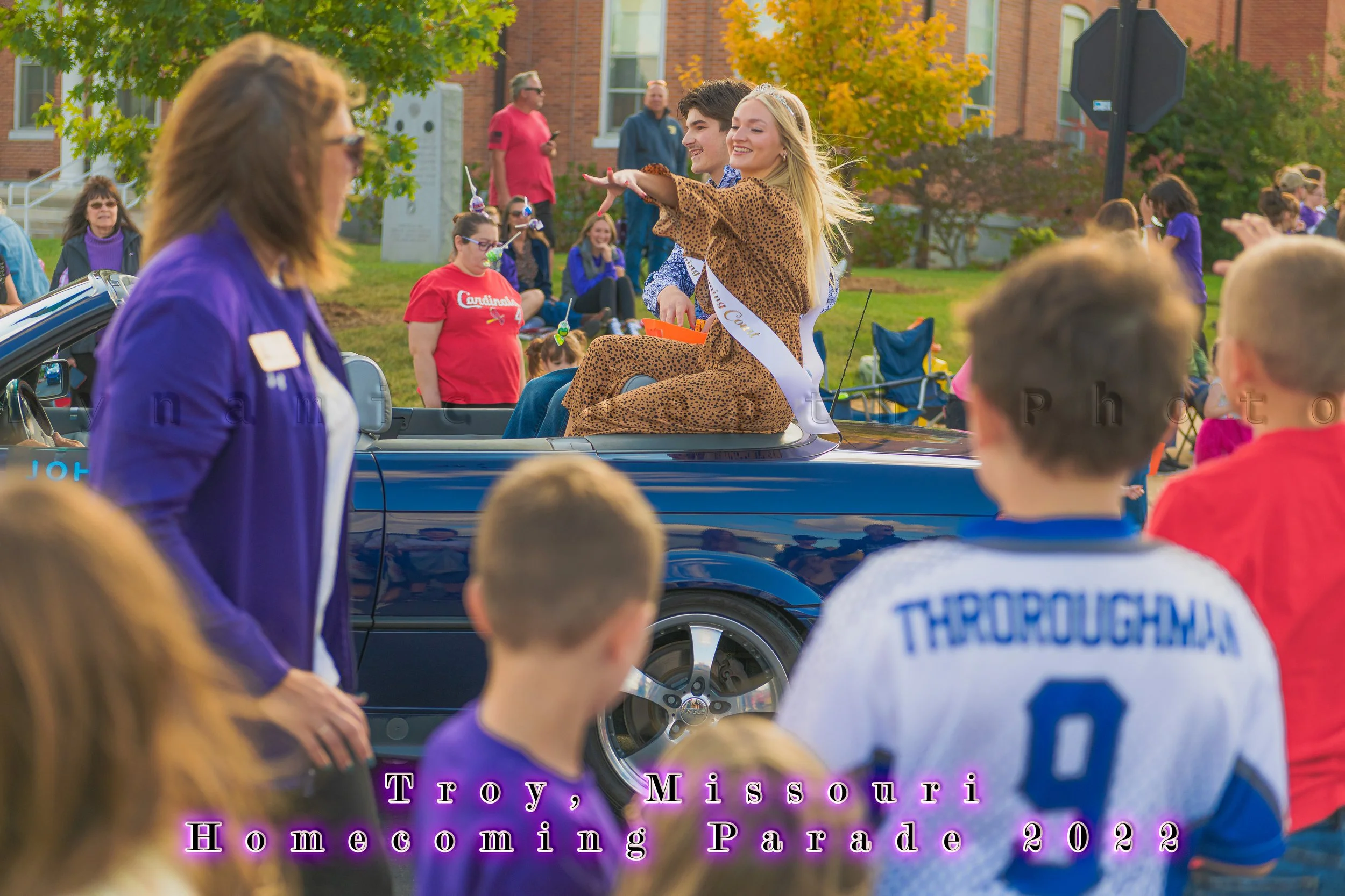 A sash-wearing passenger waves from the back of a blue vehicle during the 2022 Troy Homecoming Parade.