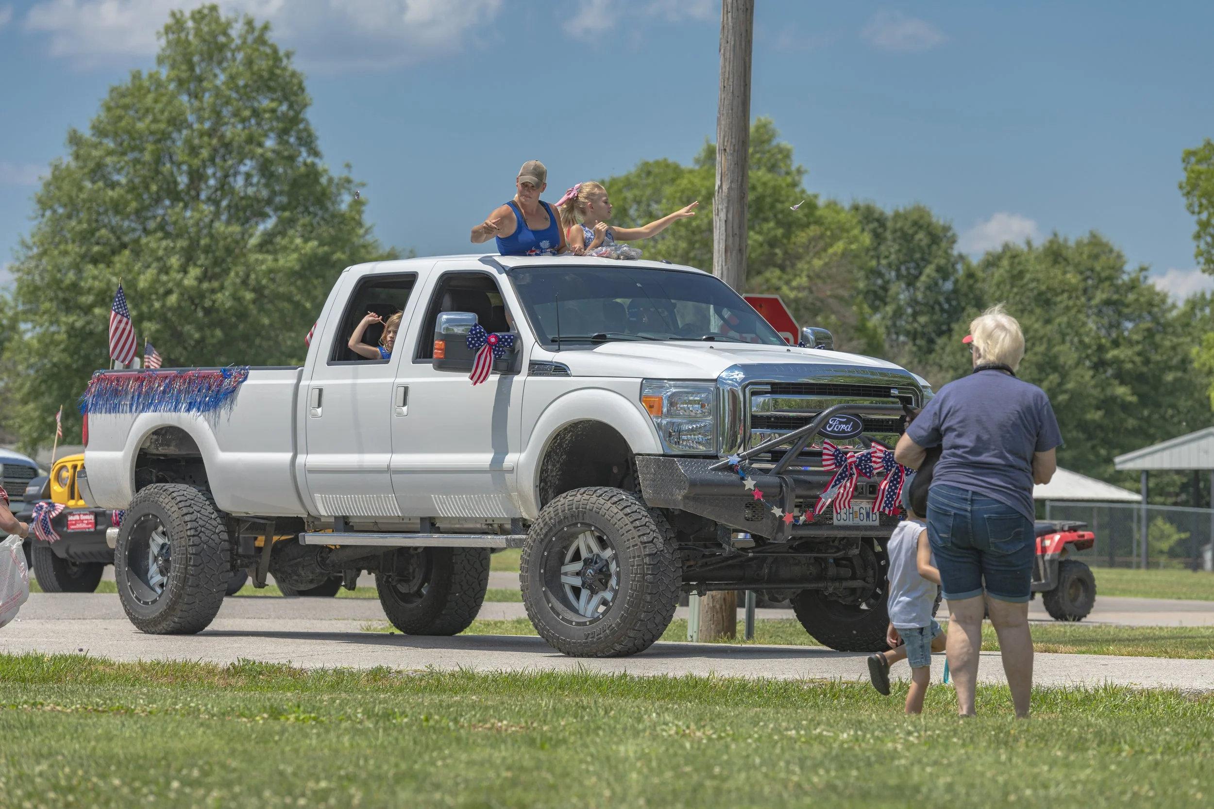 Passengers ride in a lifted silver pickup decorated with American flags during the 2022 July 4th Hawk Point Parade.