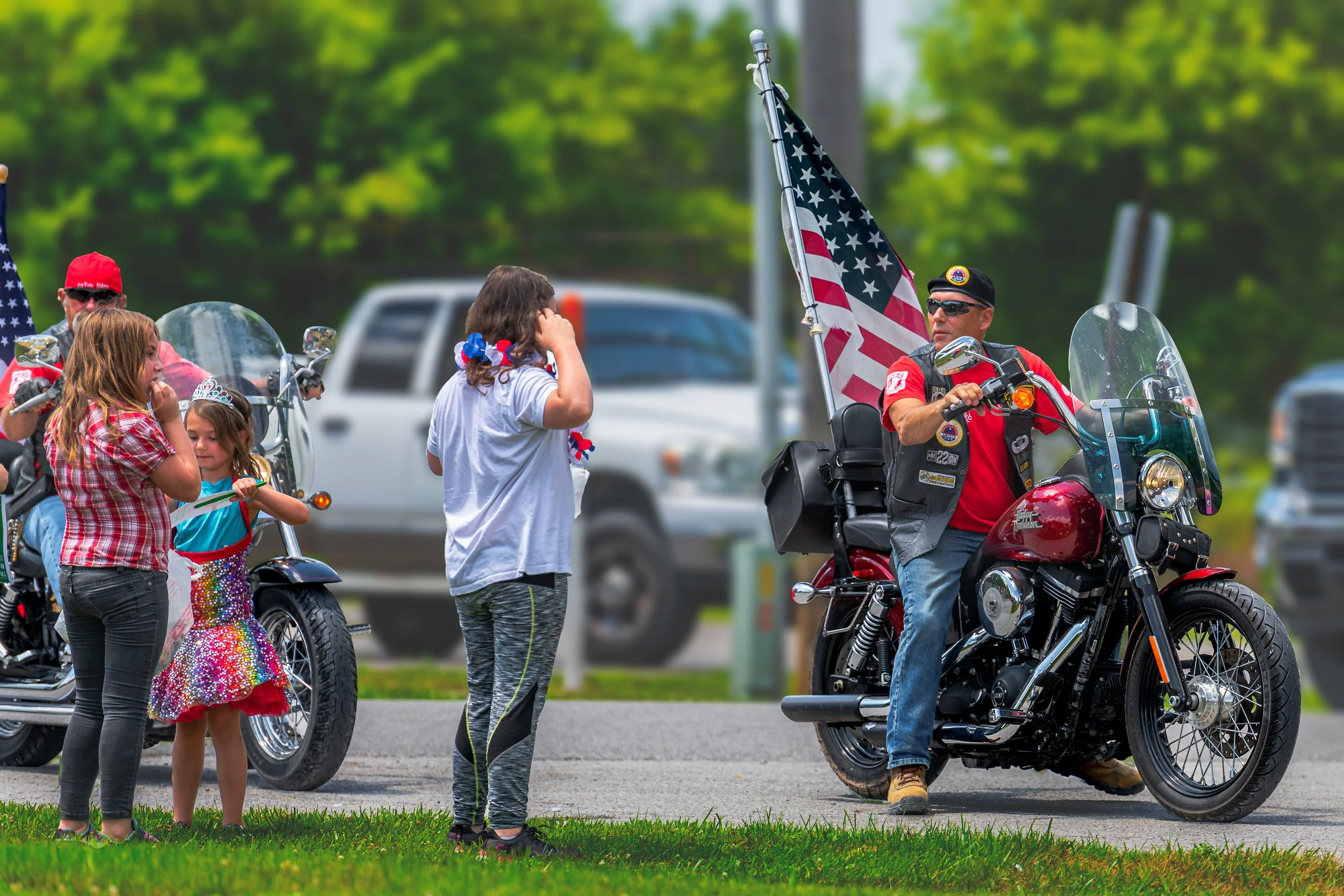 Motorcyclists pass spectators beside the road, with an American flag mounted behind a rider during the 2022 July 4th Hawk Point Parade.
