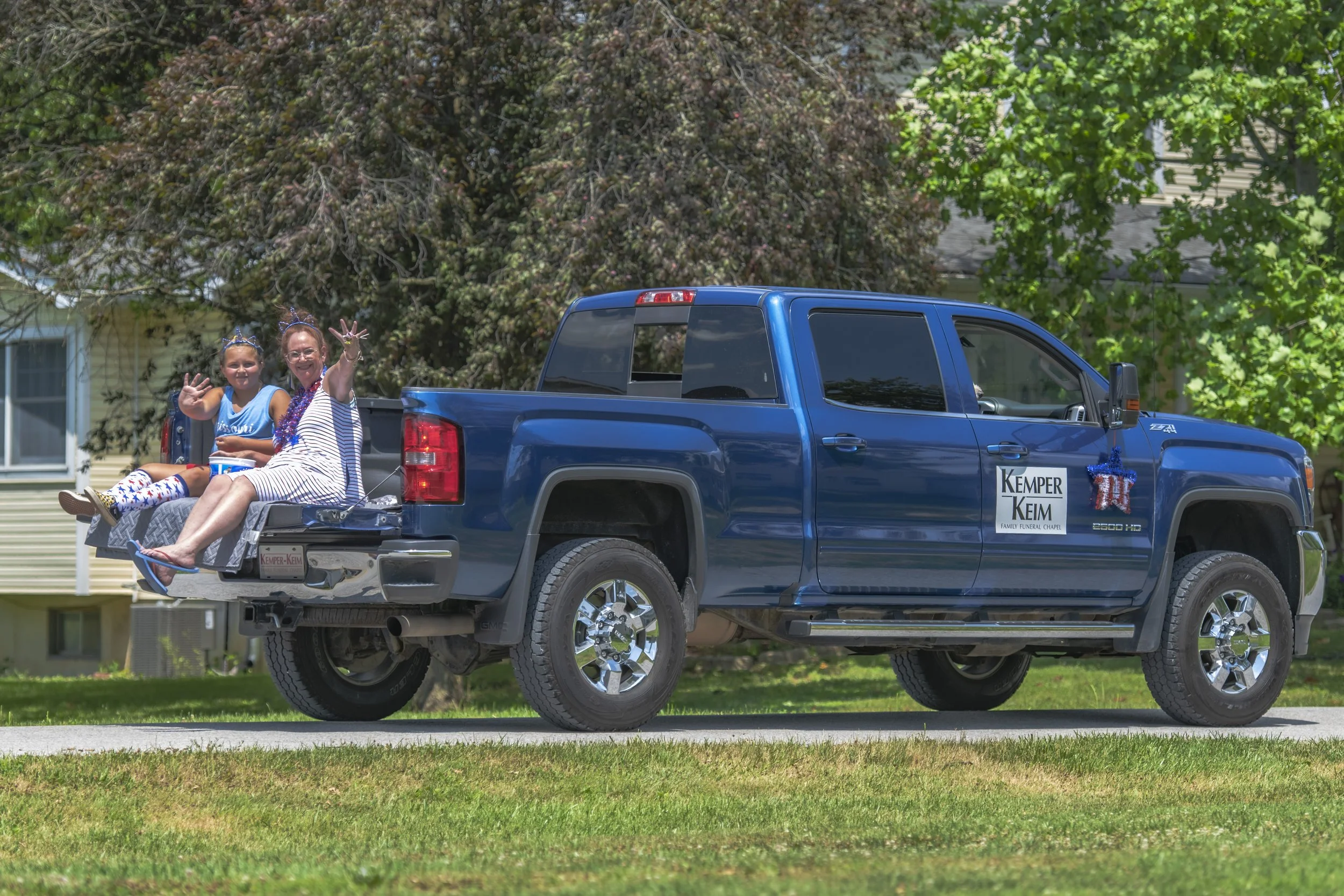 Two passengers sit on the open tailgate of a blue pickup during the 2022 July 4th Hawk Point Parade.