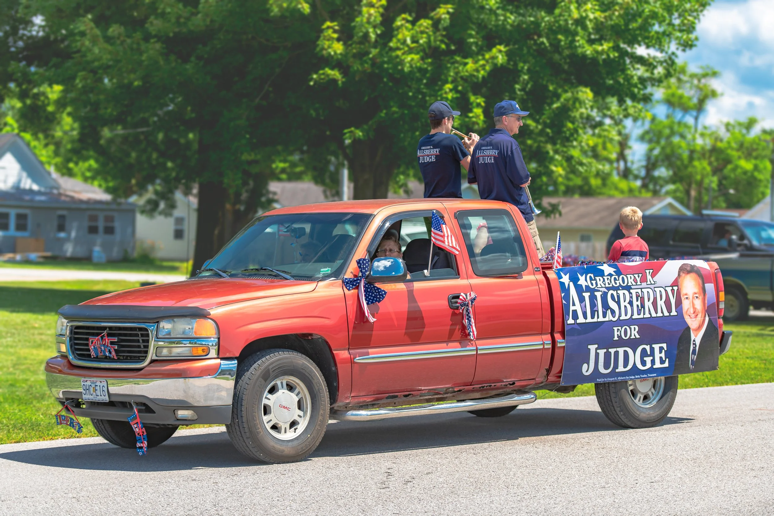Passengers ride in a red pickup carrying a large campaign banner during the 2022 July 4th Hawk Point Parade.