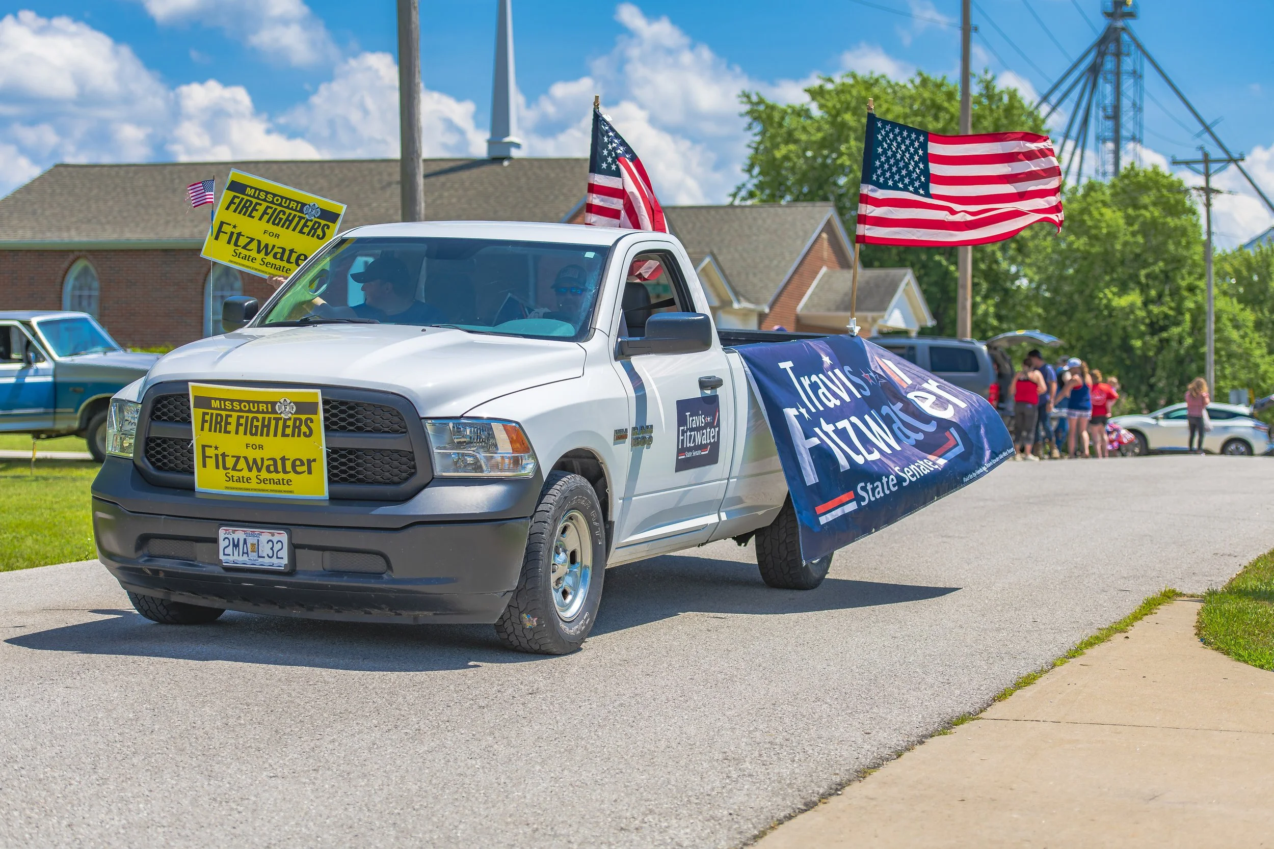 A silver pickup carries two American flags and a large campaign banner during the 2022 July 4th Hawk Point Parade.