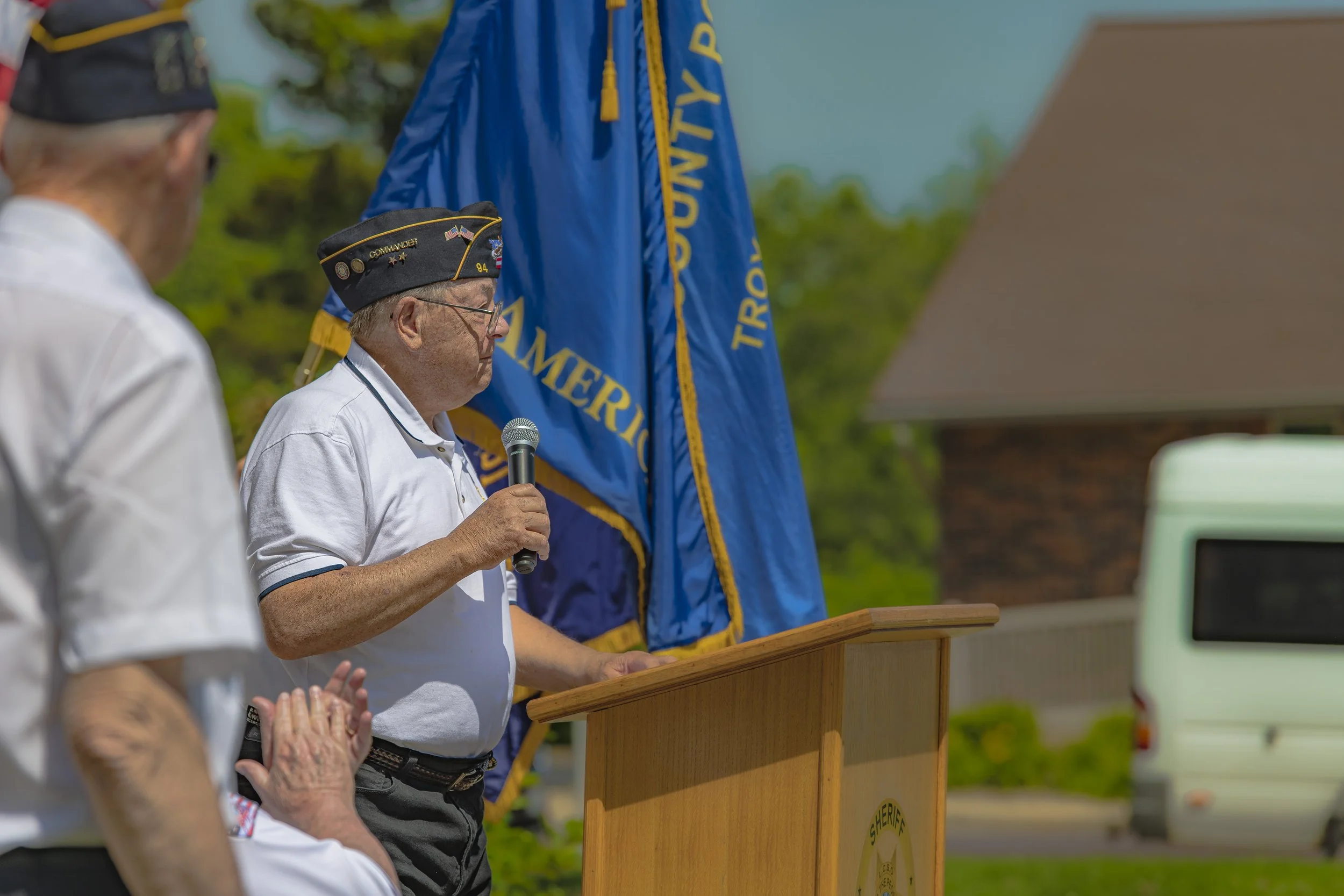 A speaker in a veterans' cap holds a microphone beside the lectern at the 2022 Lincoln County Memorial Day service.