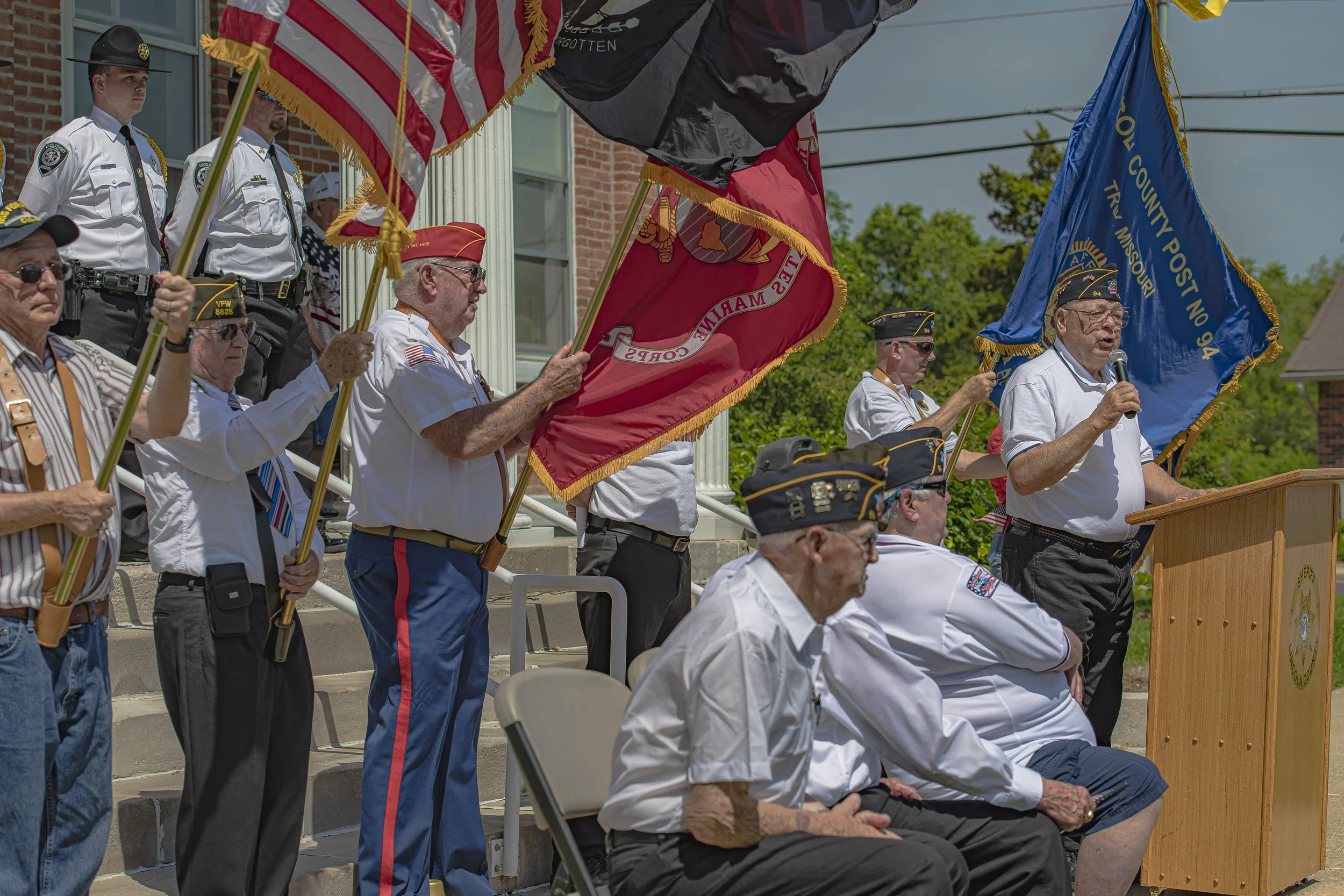 A speaker addresses the gathering beside flag bearers and seated participants at the 2022 Lincoln County Memorial Day service.