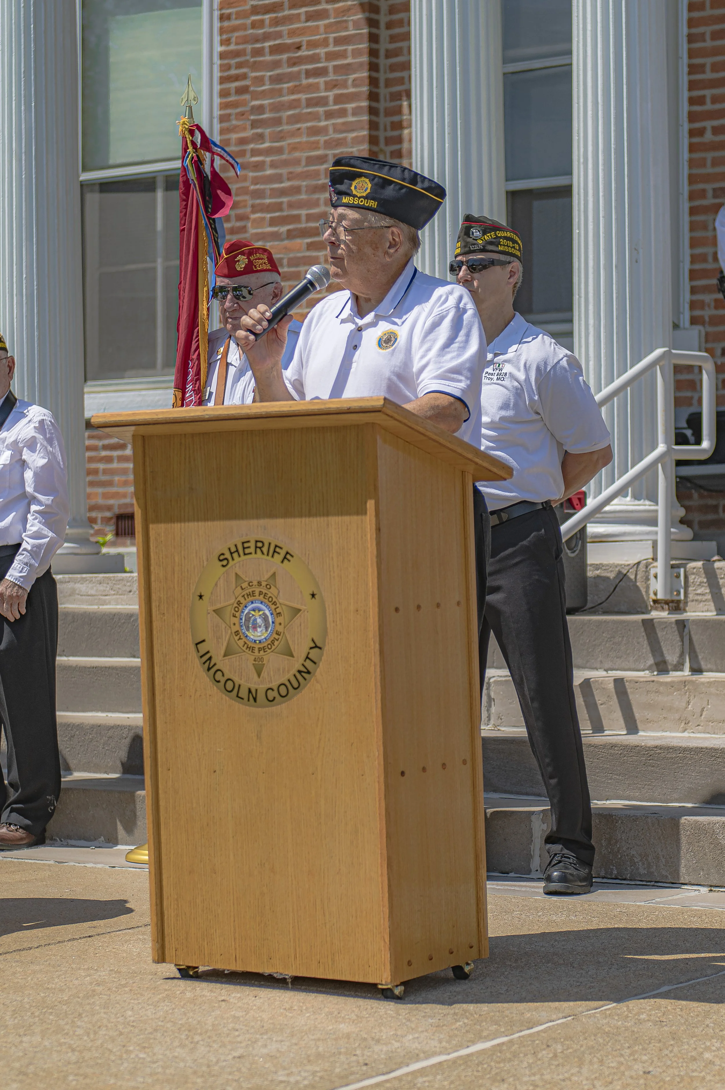 A speaker in a white polo and veterans' cap addresses the audience at the 2022 Lincoln County Memorial Day service.