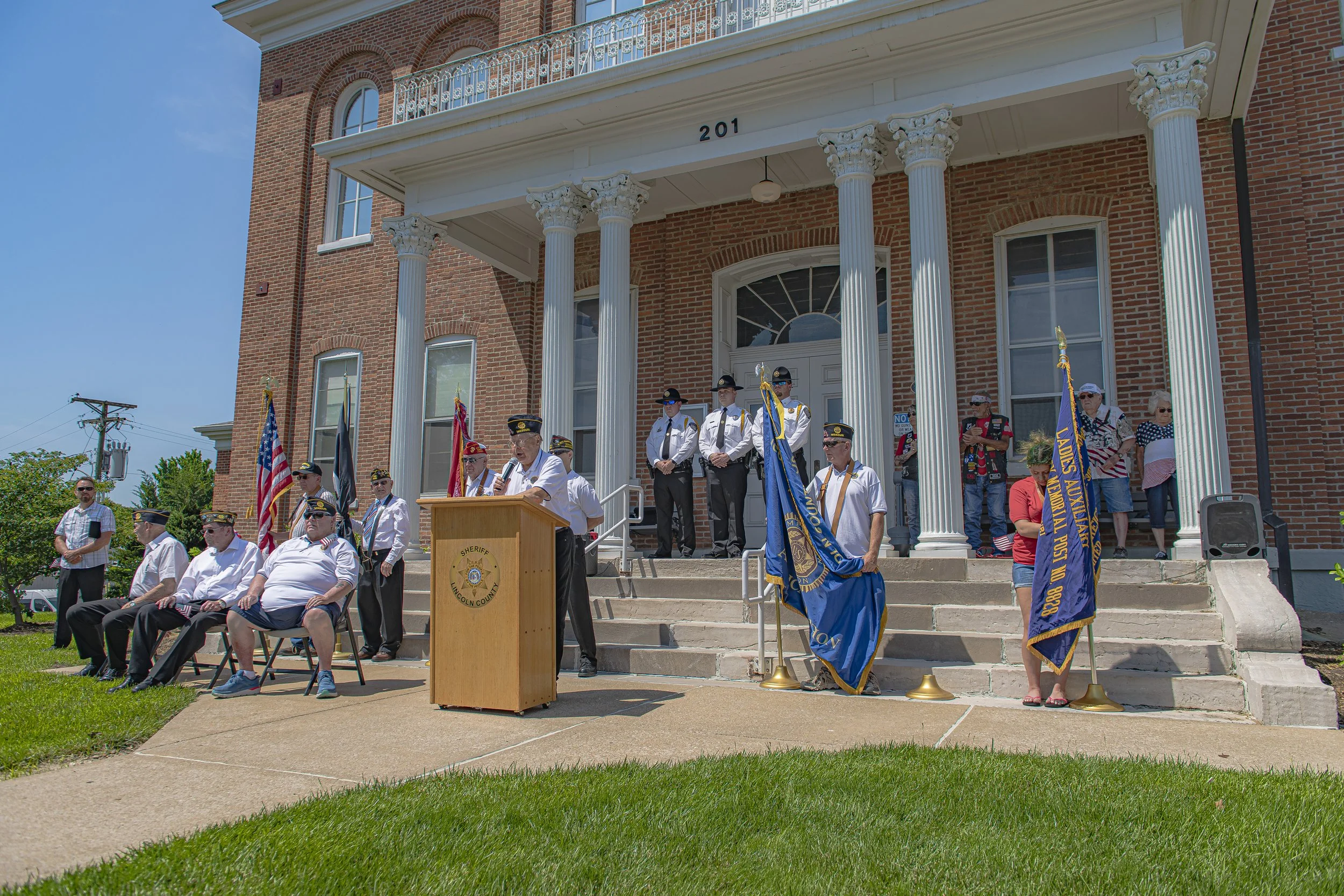 A wide view shows speakers, flags, and seated participants outside the columned entrance at the 2022 Lincoln County Memorial Day service.