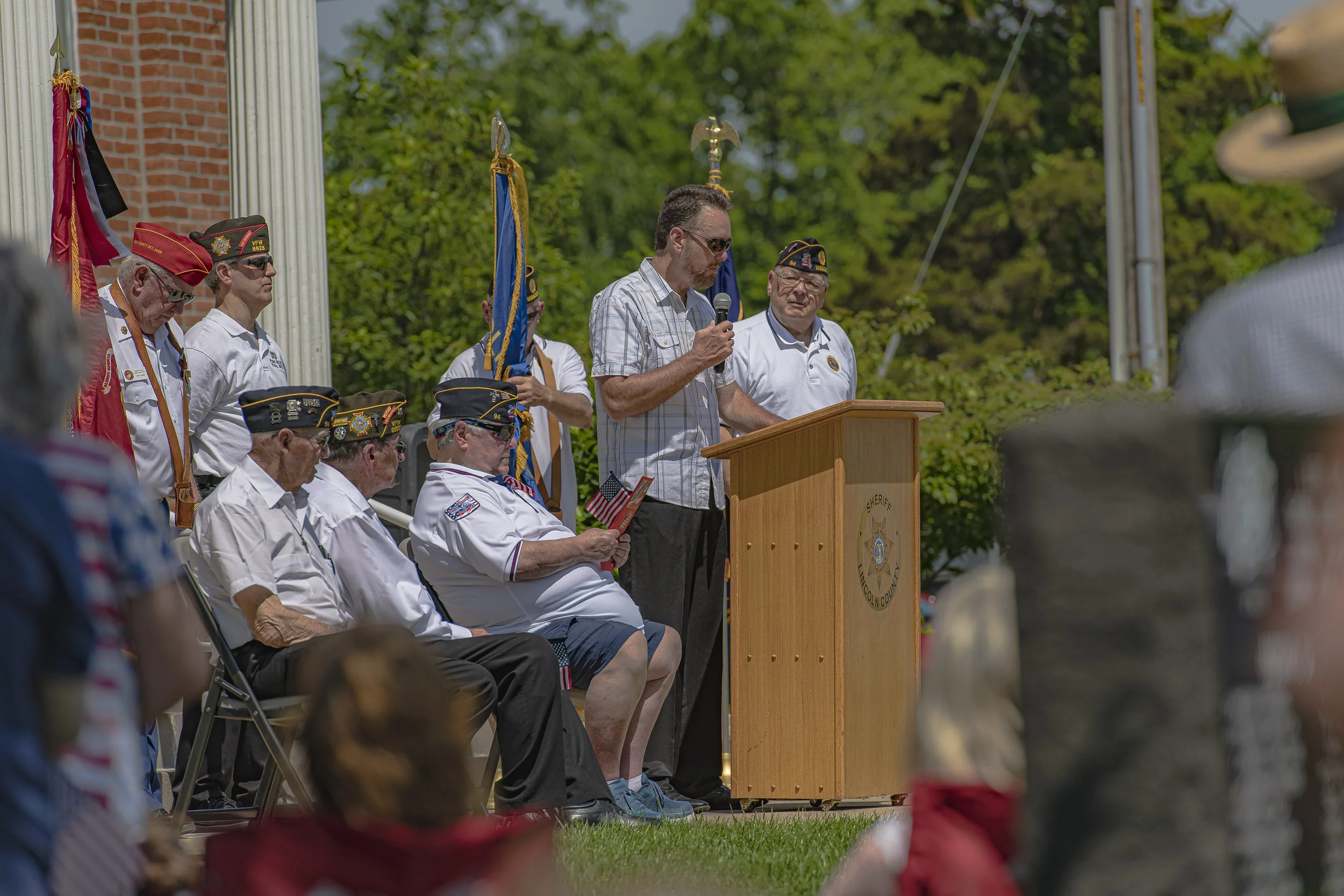 A speaker addresses the audience while participants in veterans' caps sit nearby at the 2022 Lincoln County Memorial Day service.