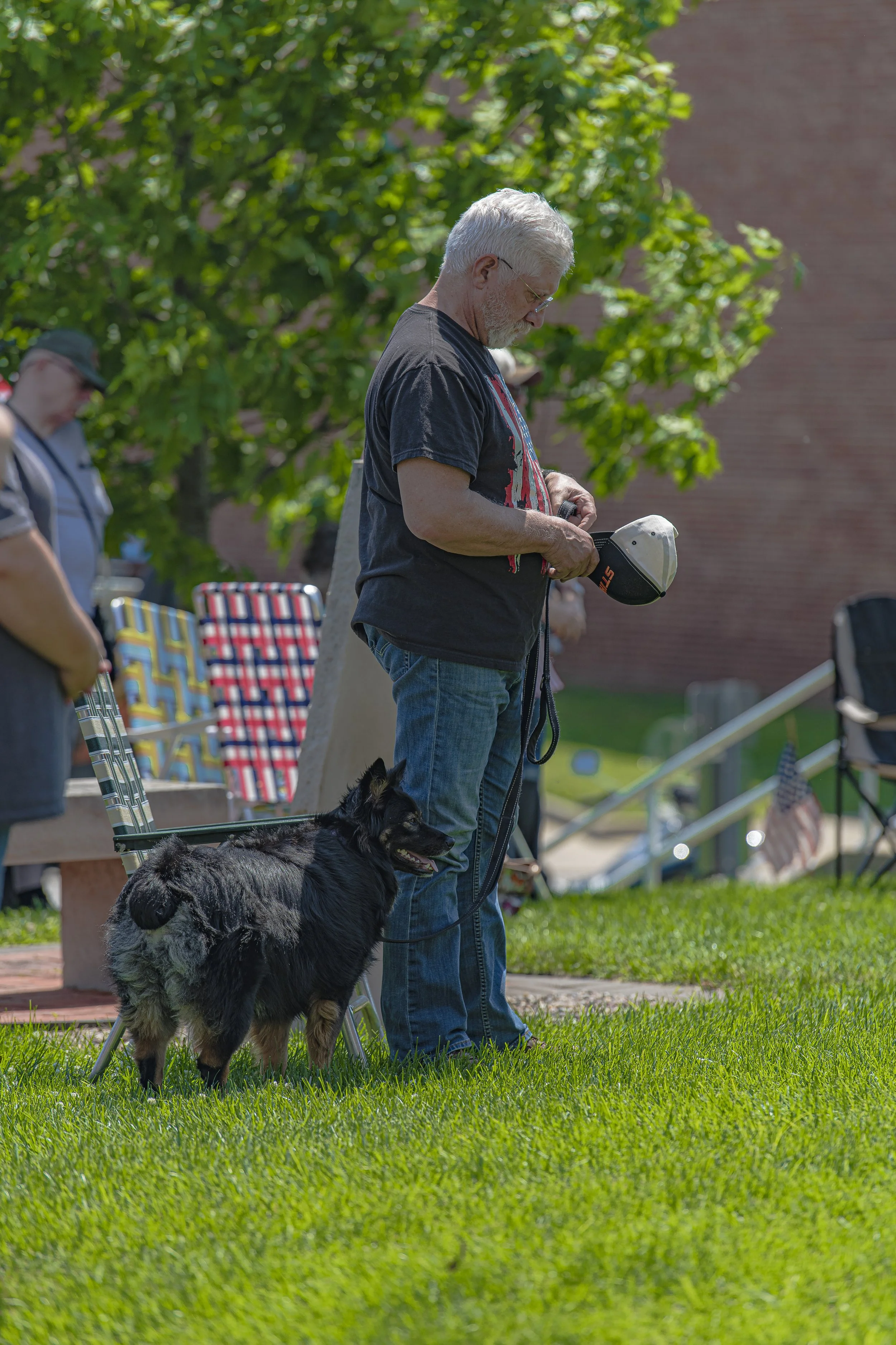 An attendee stands with a dog on a leash and holds a cap in both hands at the 2022 Lincoln County Memorial Day service.