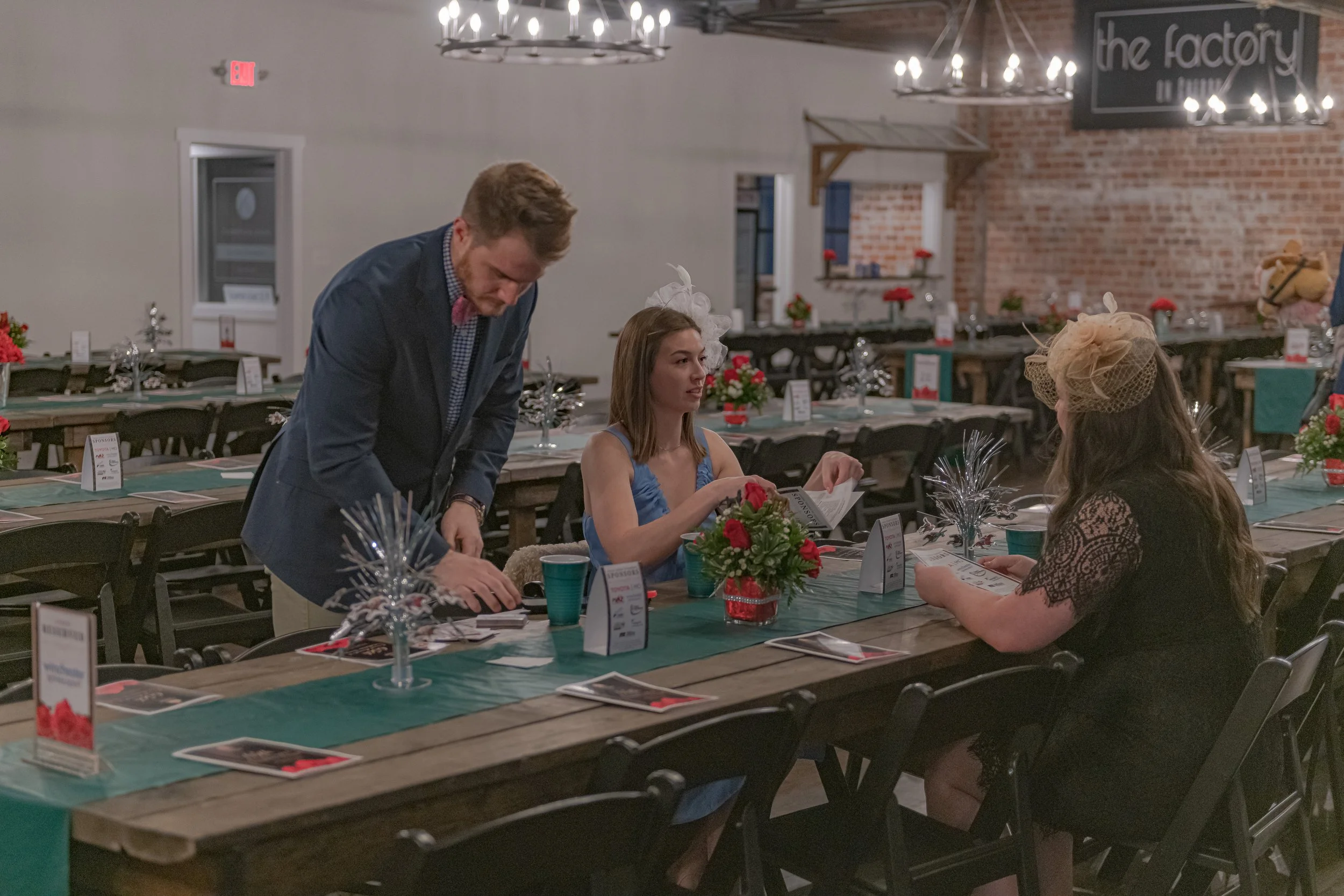 Three people gather at a banquet table with a teal runner, red flower centerpieces and papers.