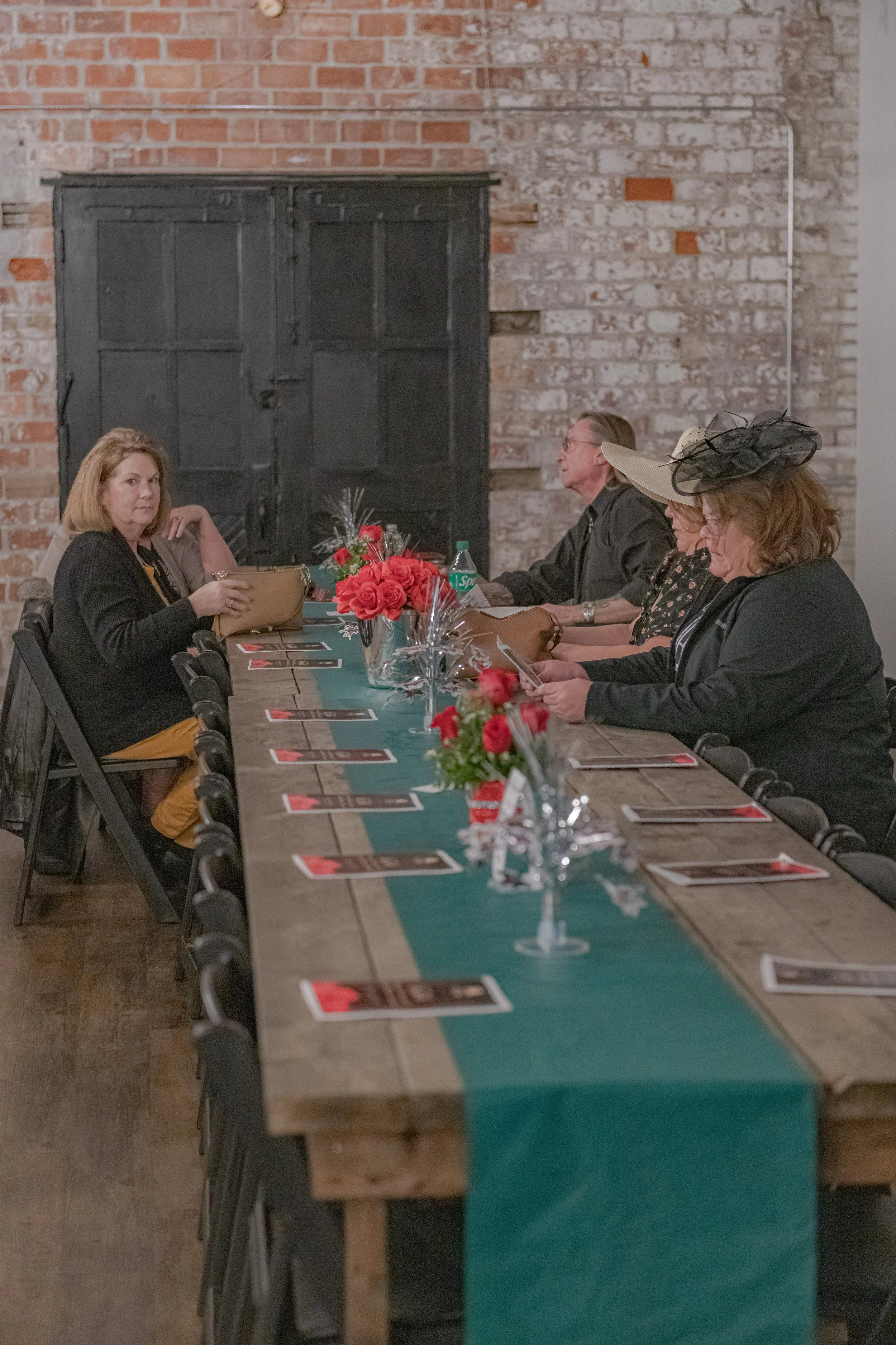 Four people sit at a long banquet table decorated with a teal runner, red flowers and silver ornaments.