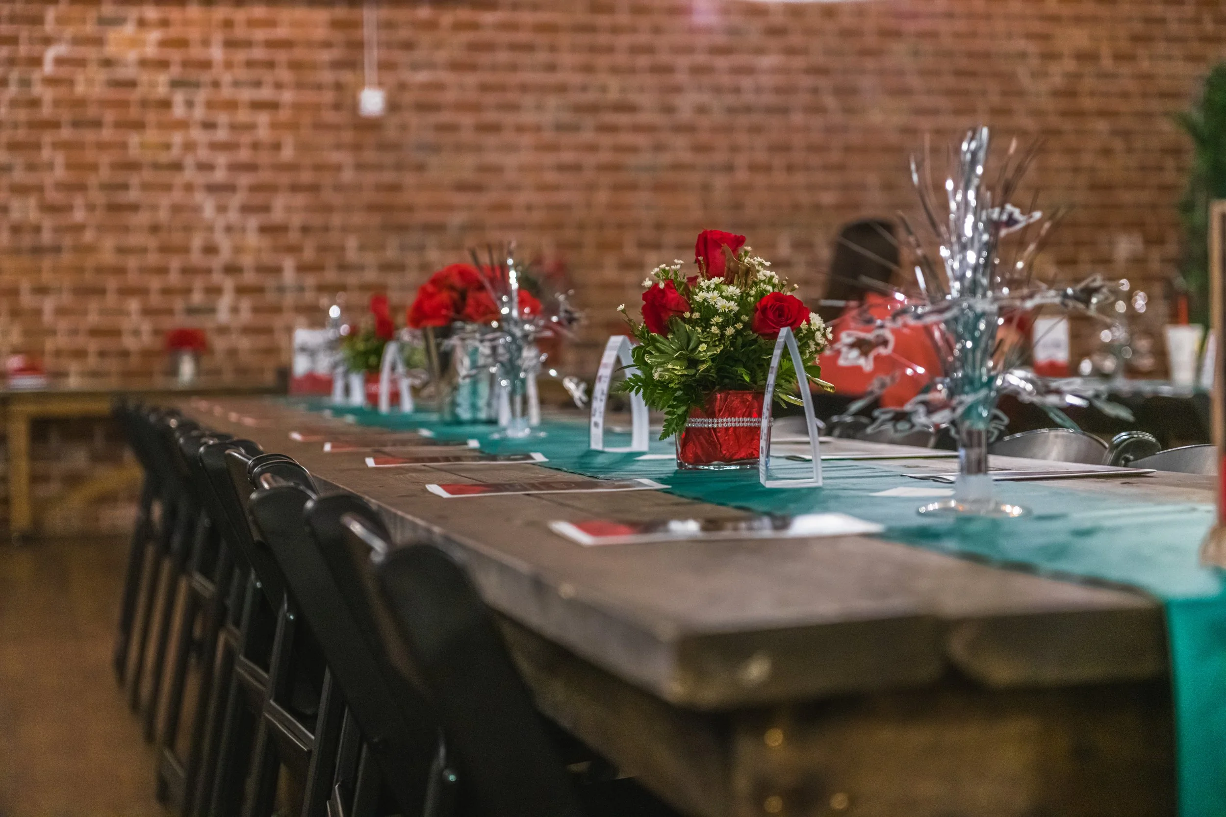 Red flower centerpieces, silver decorations and printed programs line a long wooden banquet table.