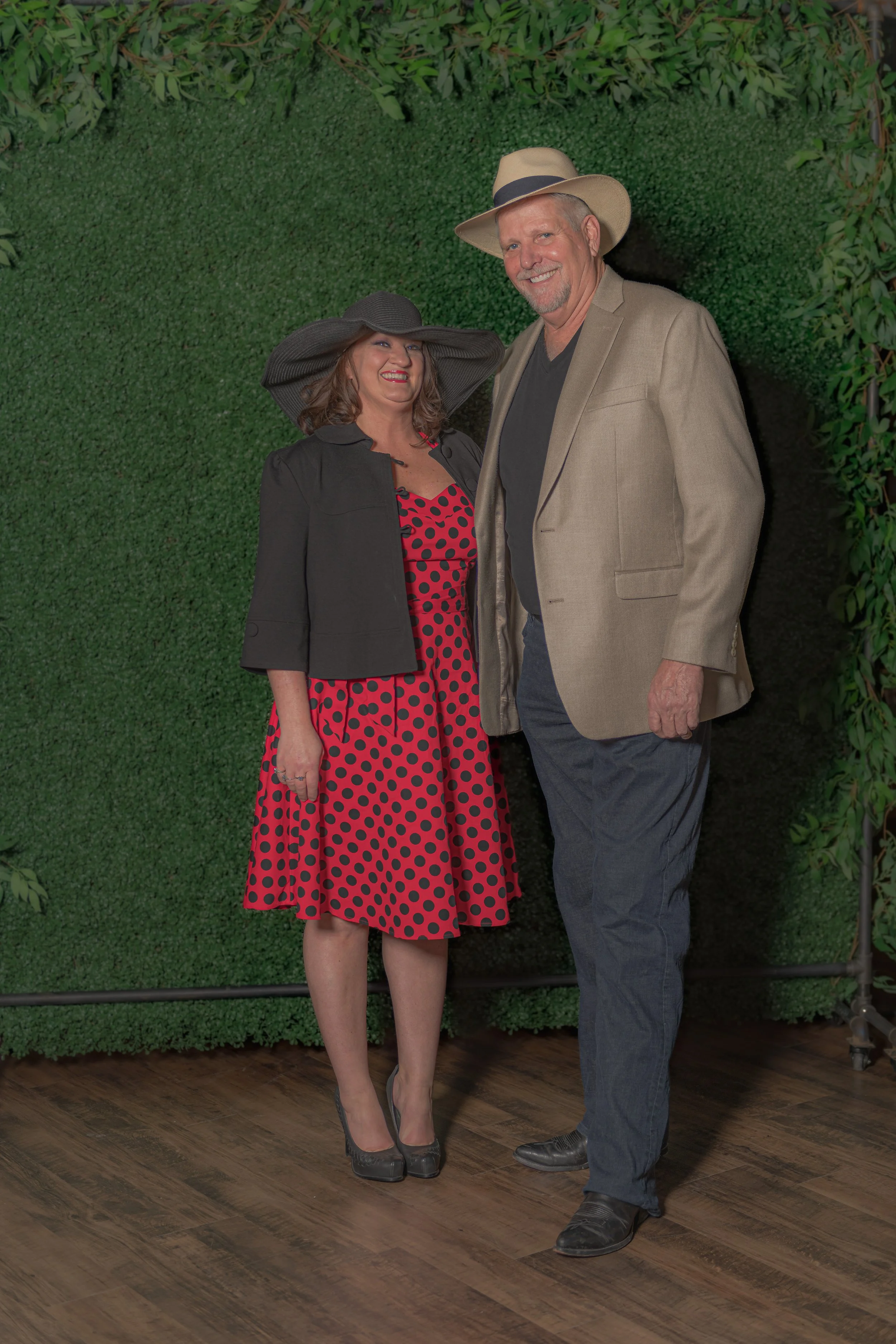 Two people in wide-brimmed hats pose against a green foliage backdrop, one in a red patterned dress and one in a beige jacket.