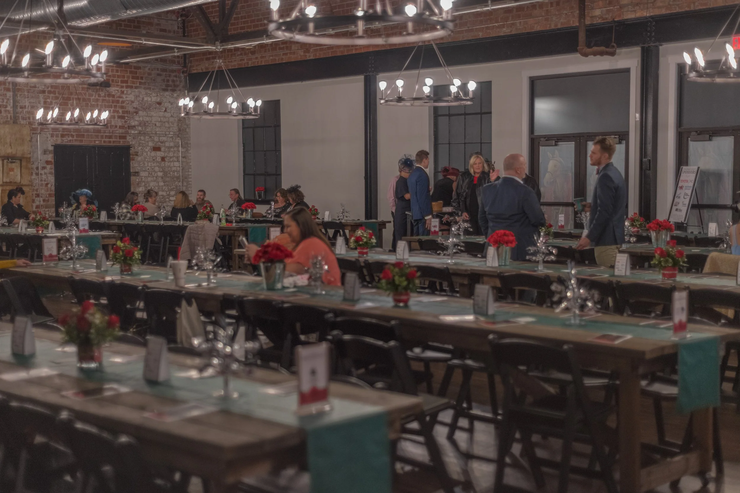 People sit and stand among rows of decorated wooden tables beneath chandeliers in a brick-walled banquet room.