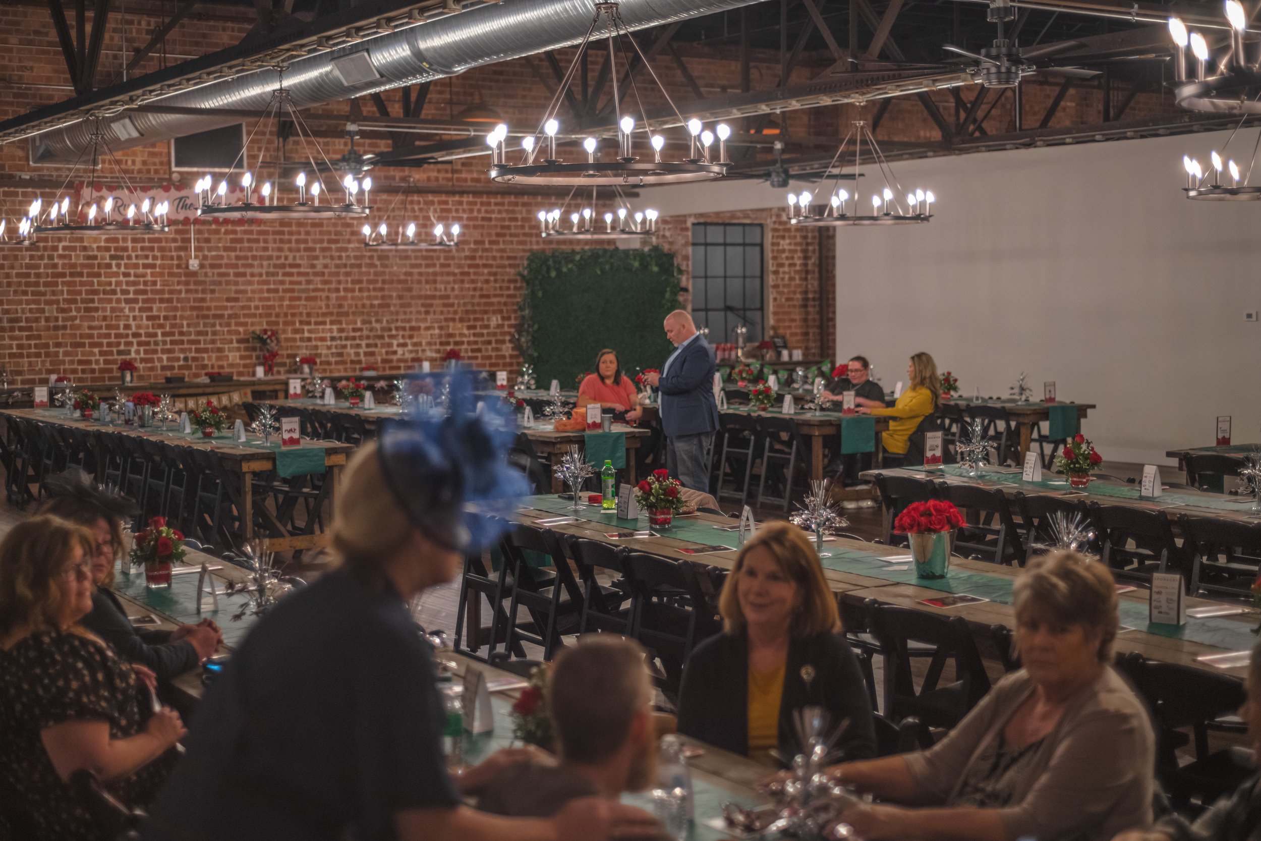 People gather around banquet tables beneath chandeliers, with a blue feathered headpiece in the foreground.