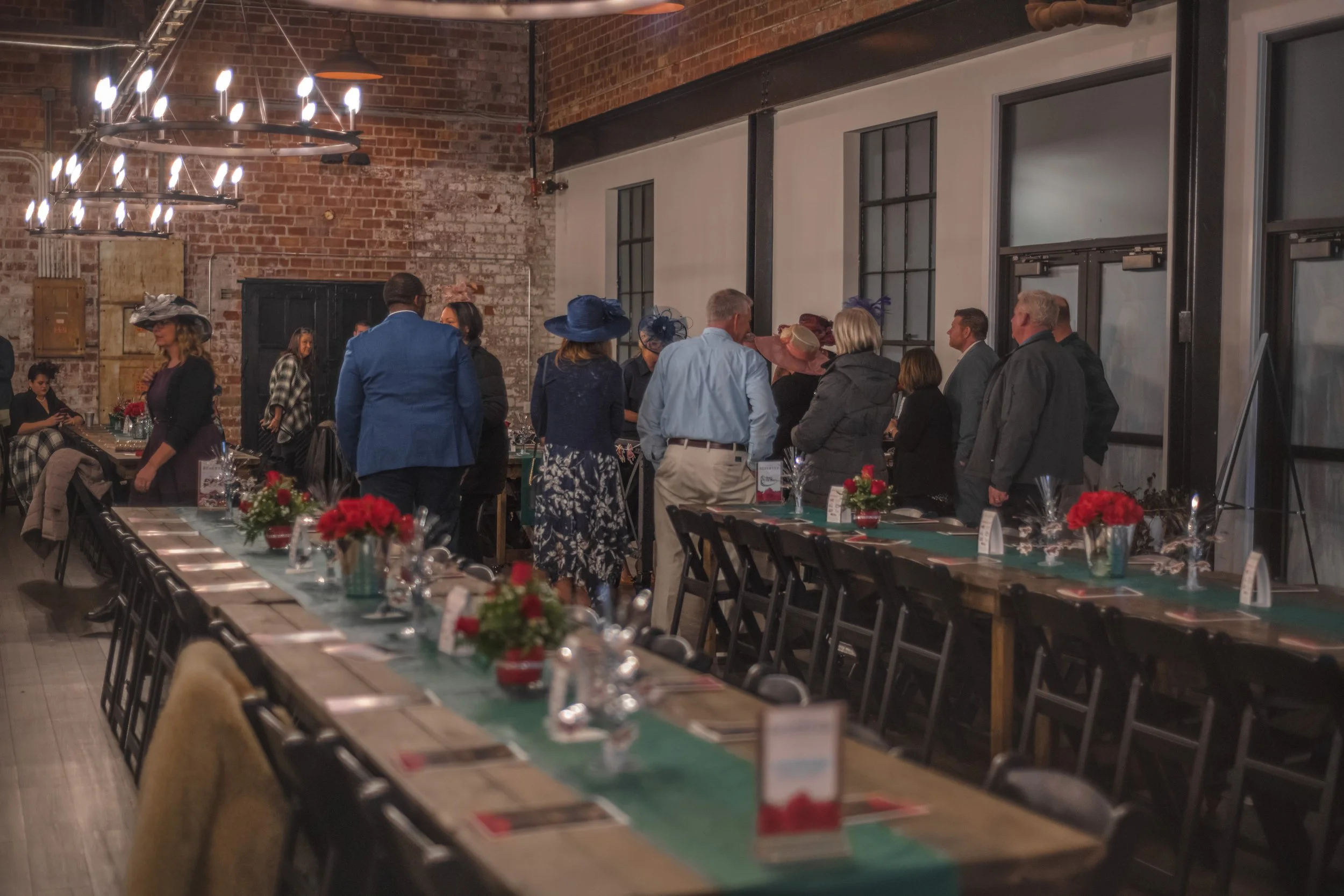 People stand near tall windows beside banquet tables set with red flowers and teal runners.