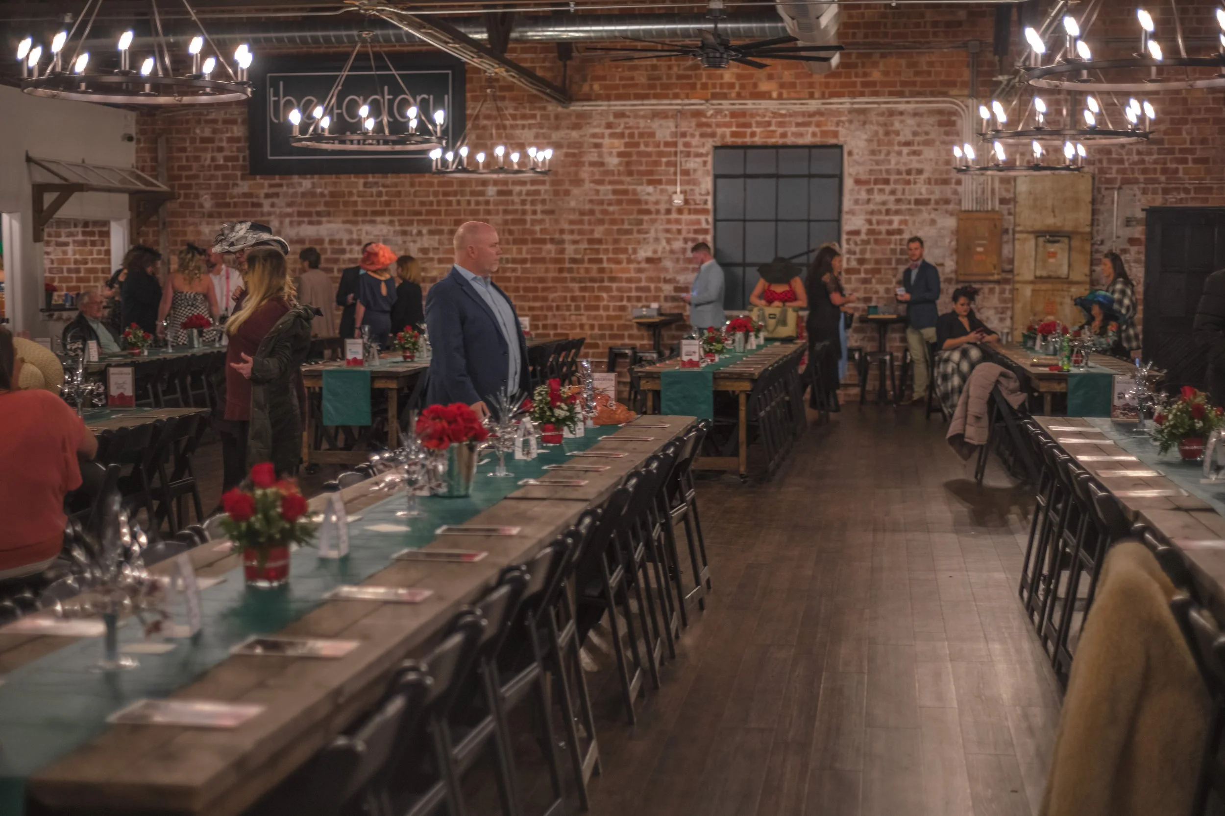 A wide aisle runs between banquet tables toward people standing beneath a “the factory” sign on a brick wall.