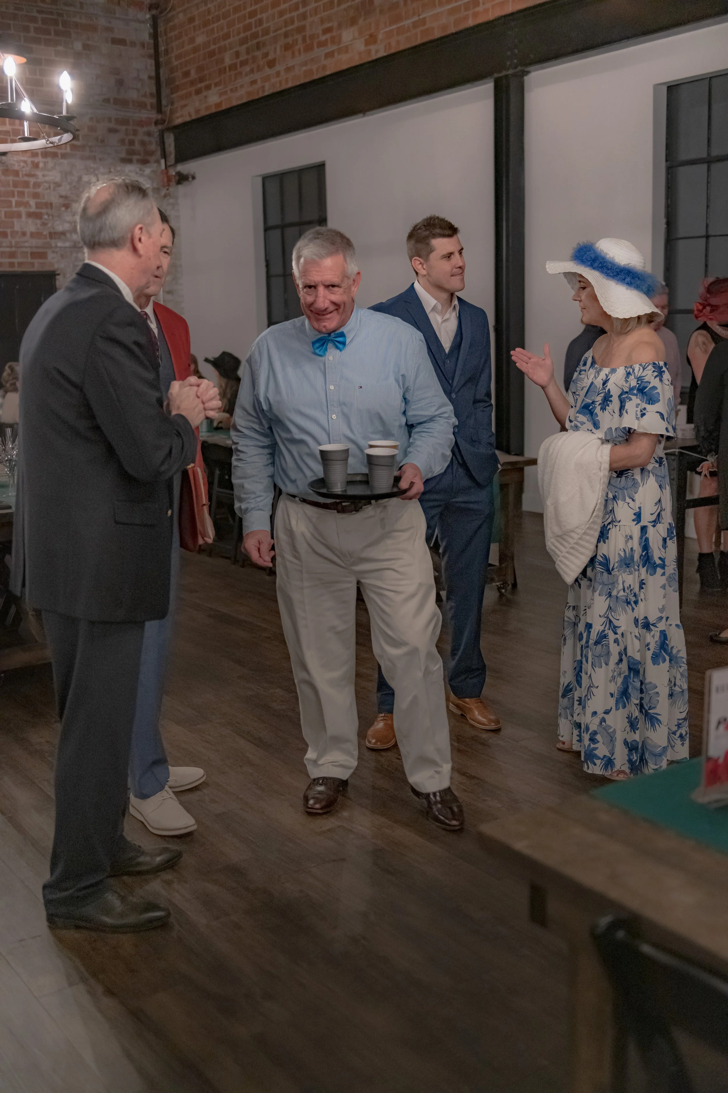A person wearing a blue bow tie carries a tray of cups among people standing beside a banquet table.