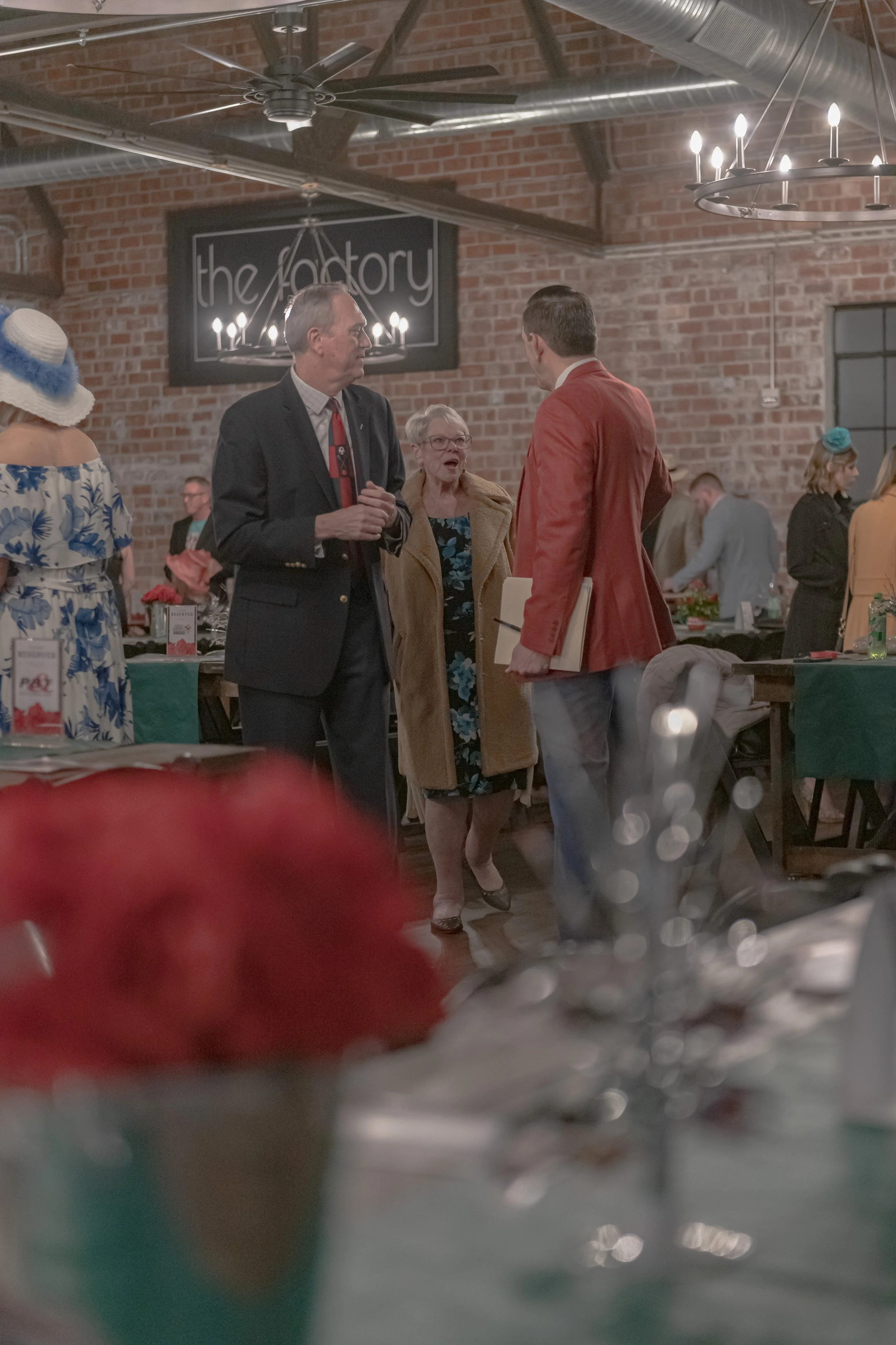 Three people stand behind a blurred red-flower centerpiece, including someone in a red blazer facing away.