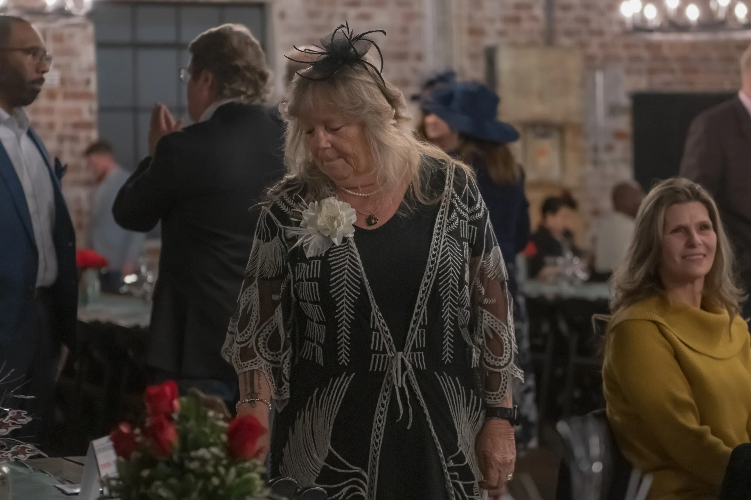 A person in a black outfit with a patterned sheer overlay and white flower corsage stands beside a banquet table.