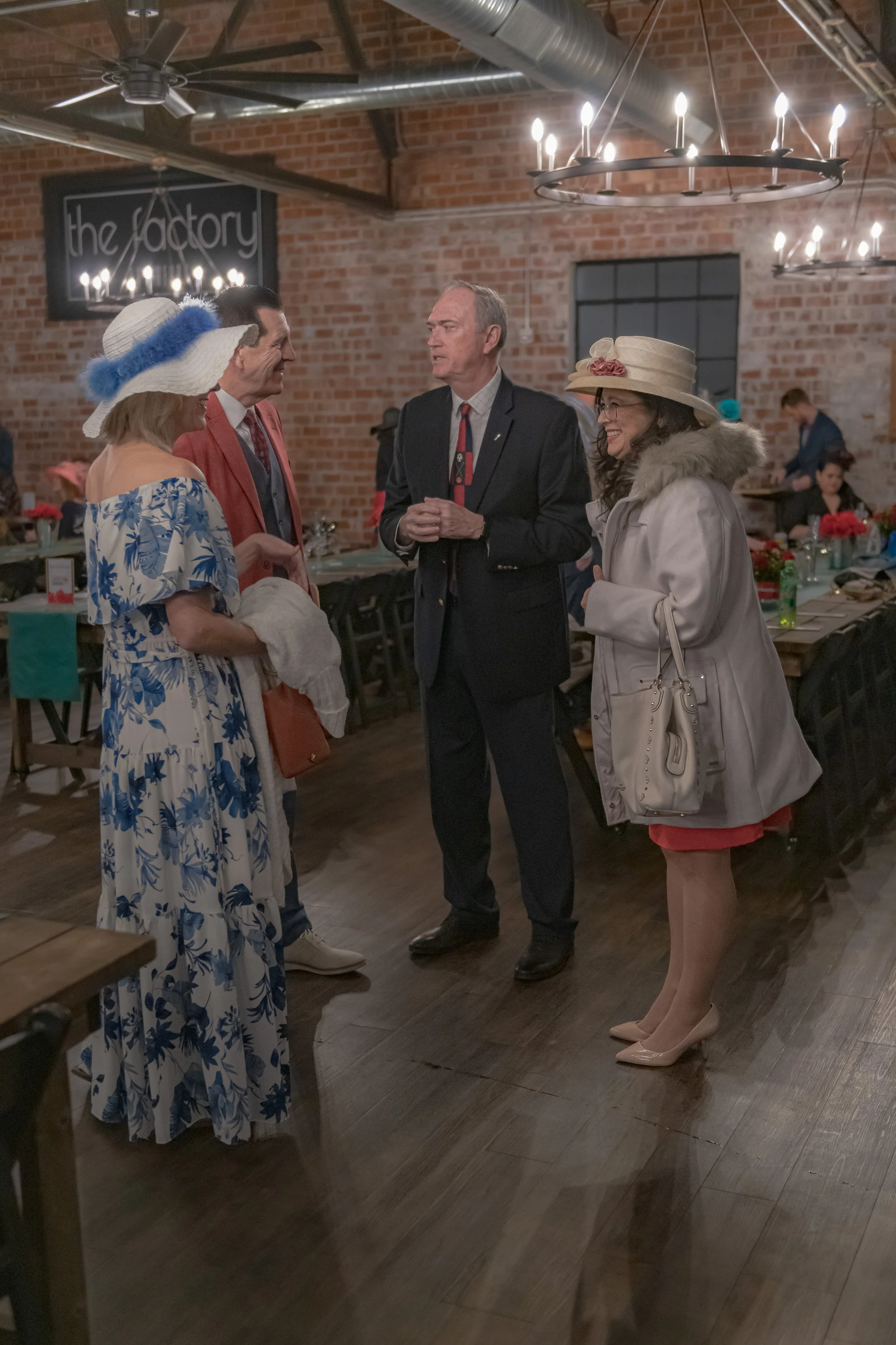 Four people stand together between banquet tables, including two in wide-brimmed hats and one in a red blazer.