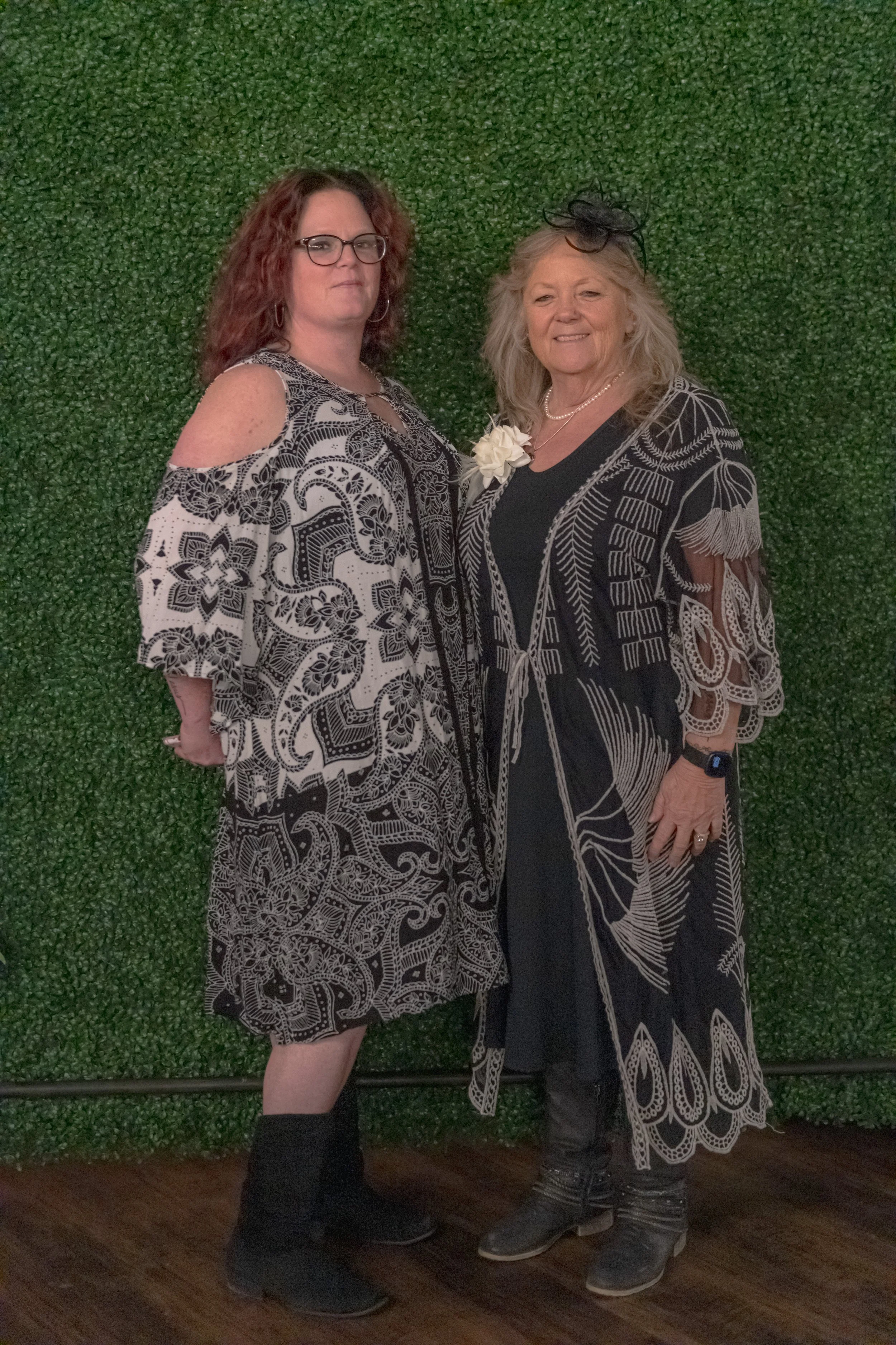 Two people in black-and-white patterned outfits pose side by side against a green foliage backdrop.