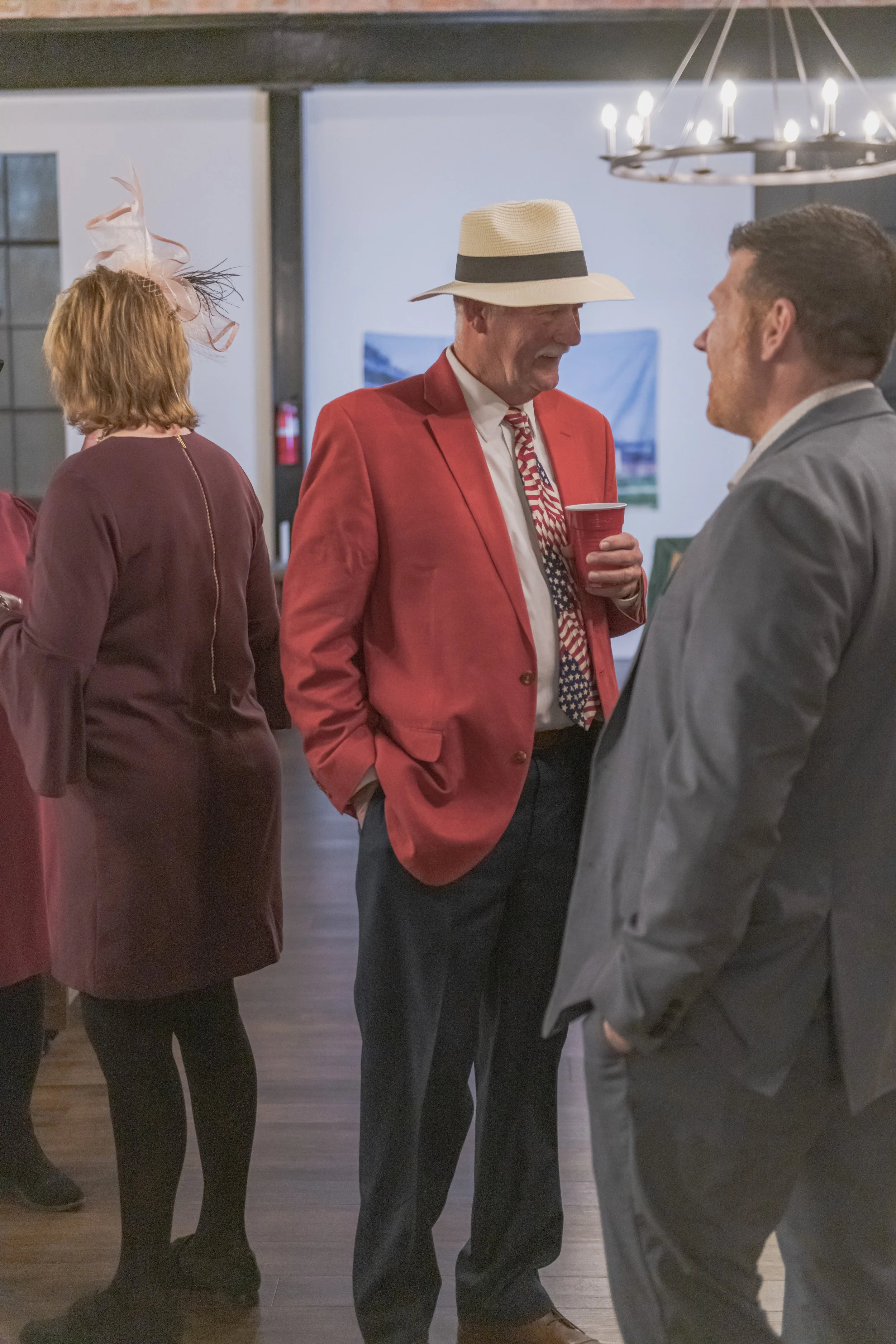 A person in a red blazer and light-colored hat faces someone in a gray suit near a banquet-room doorway.