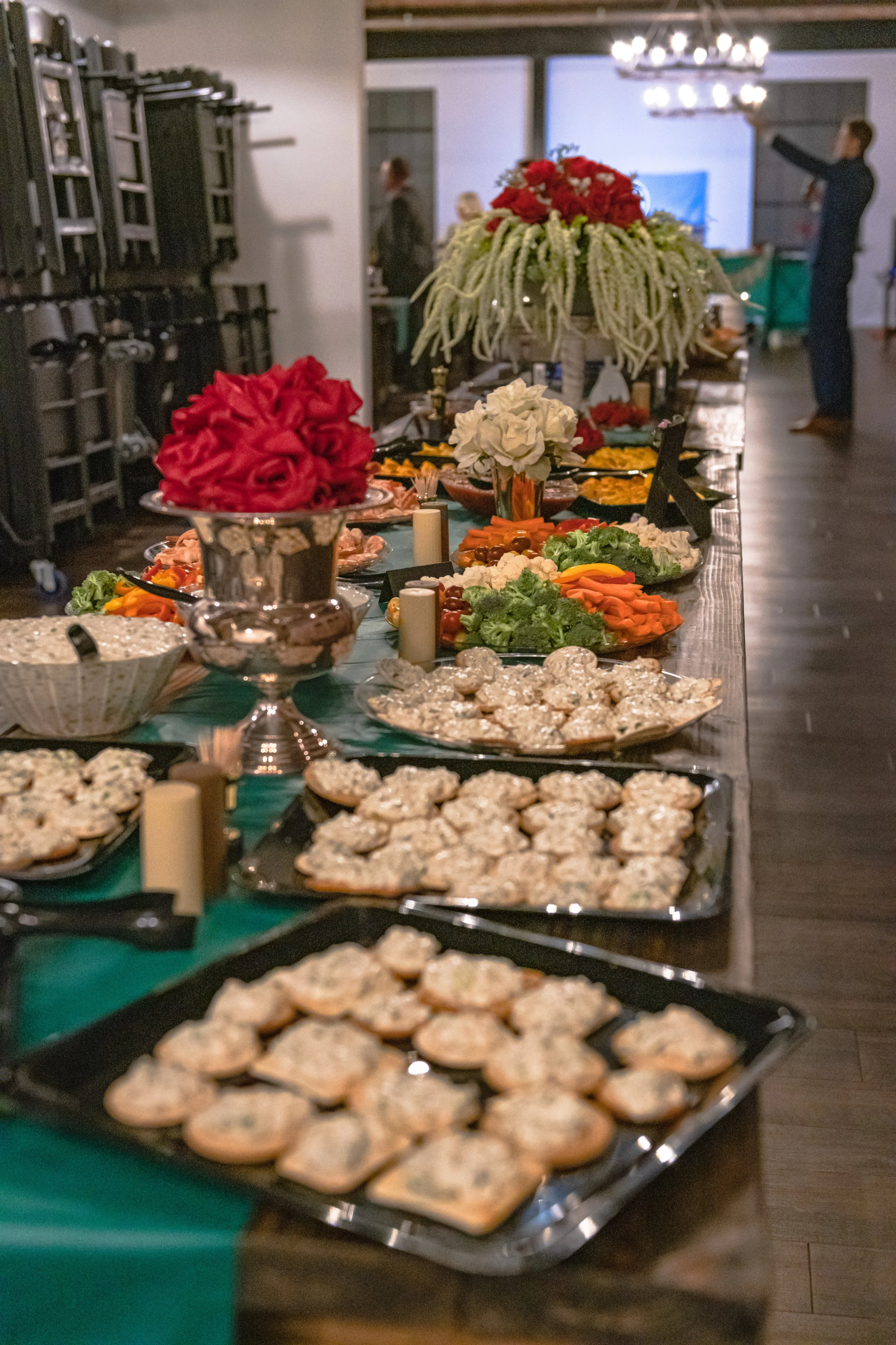 A long buffet table holds trays of bite-size food, vegetables and red flower arrangements.