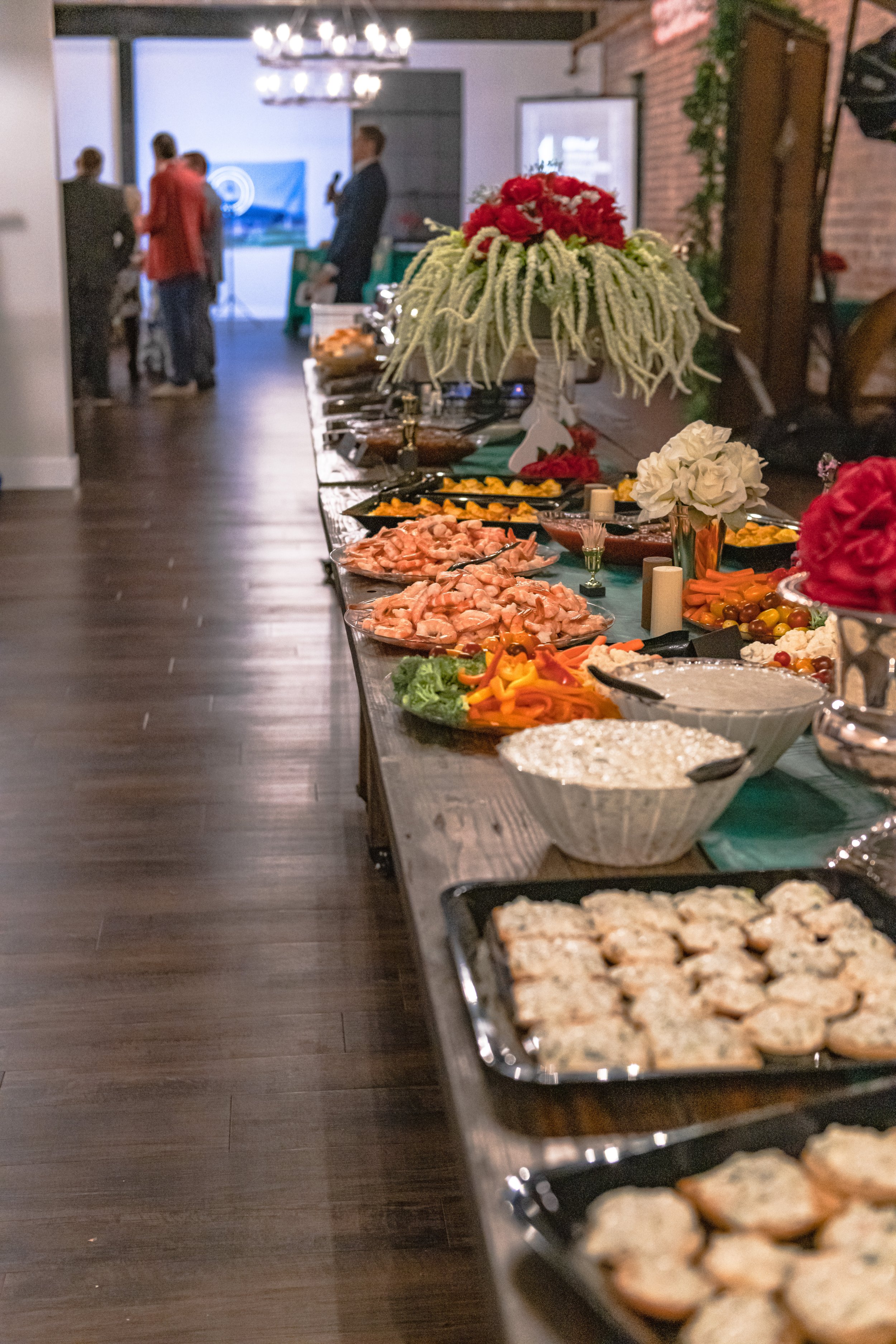 Platters of food, serving bowls and tall floral arrangements line a buffet table beside a wide aisle.