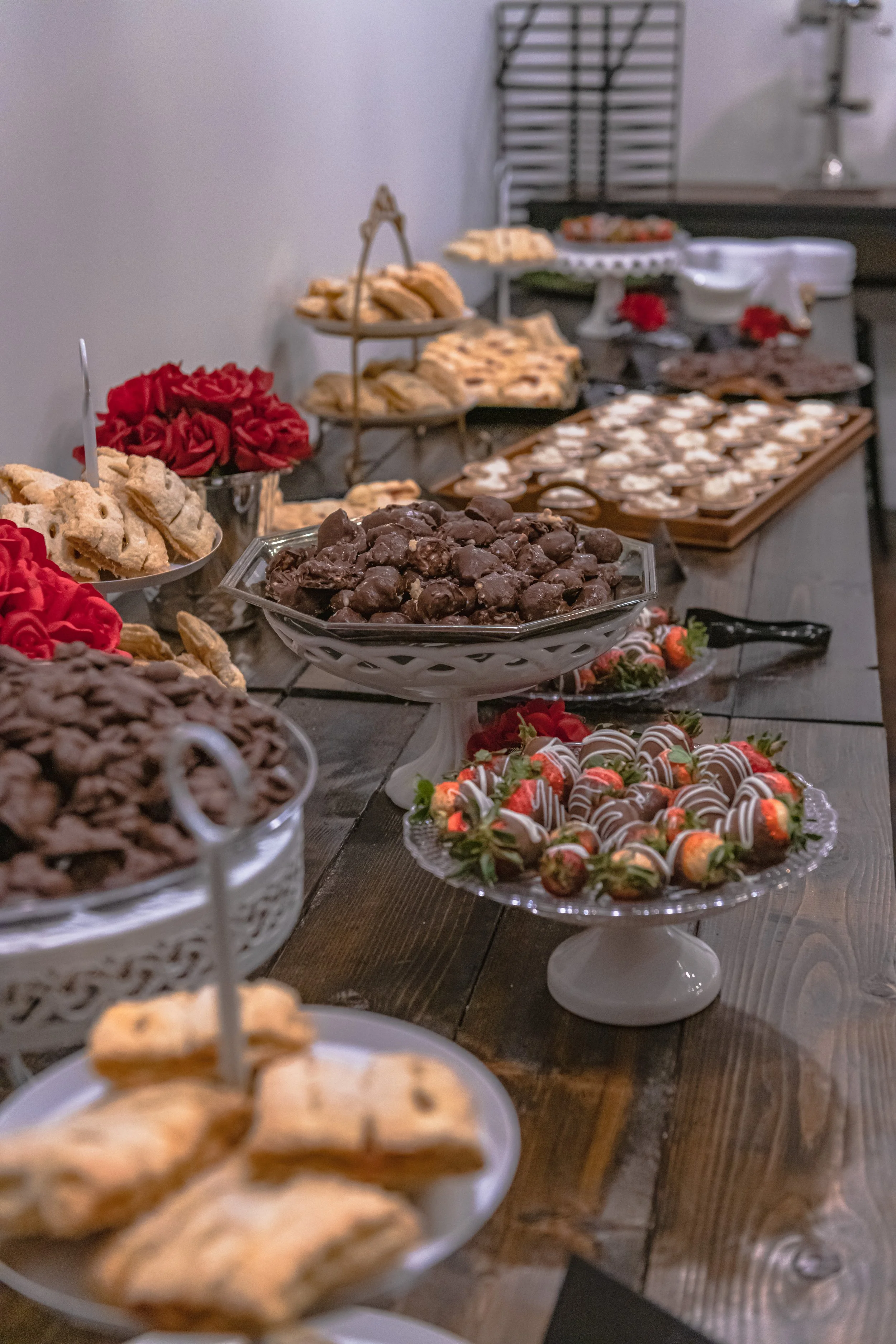 A dessert table holds chocolate-dipped strawberries, pastries and trays of small sweets beside red flowers.