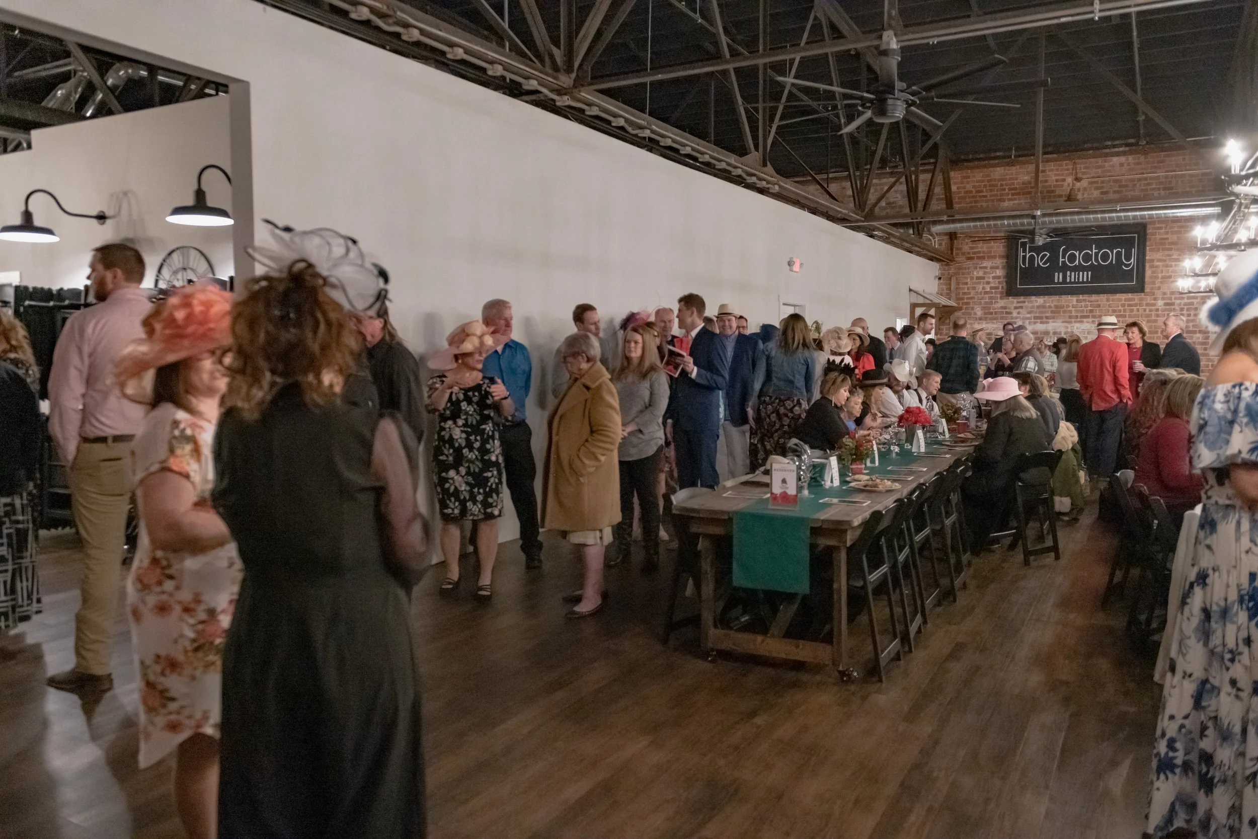 A crowd stands and sits around long banquet tables in a room with exposed ceiling beams and brick walls.