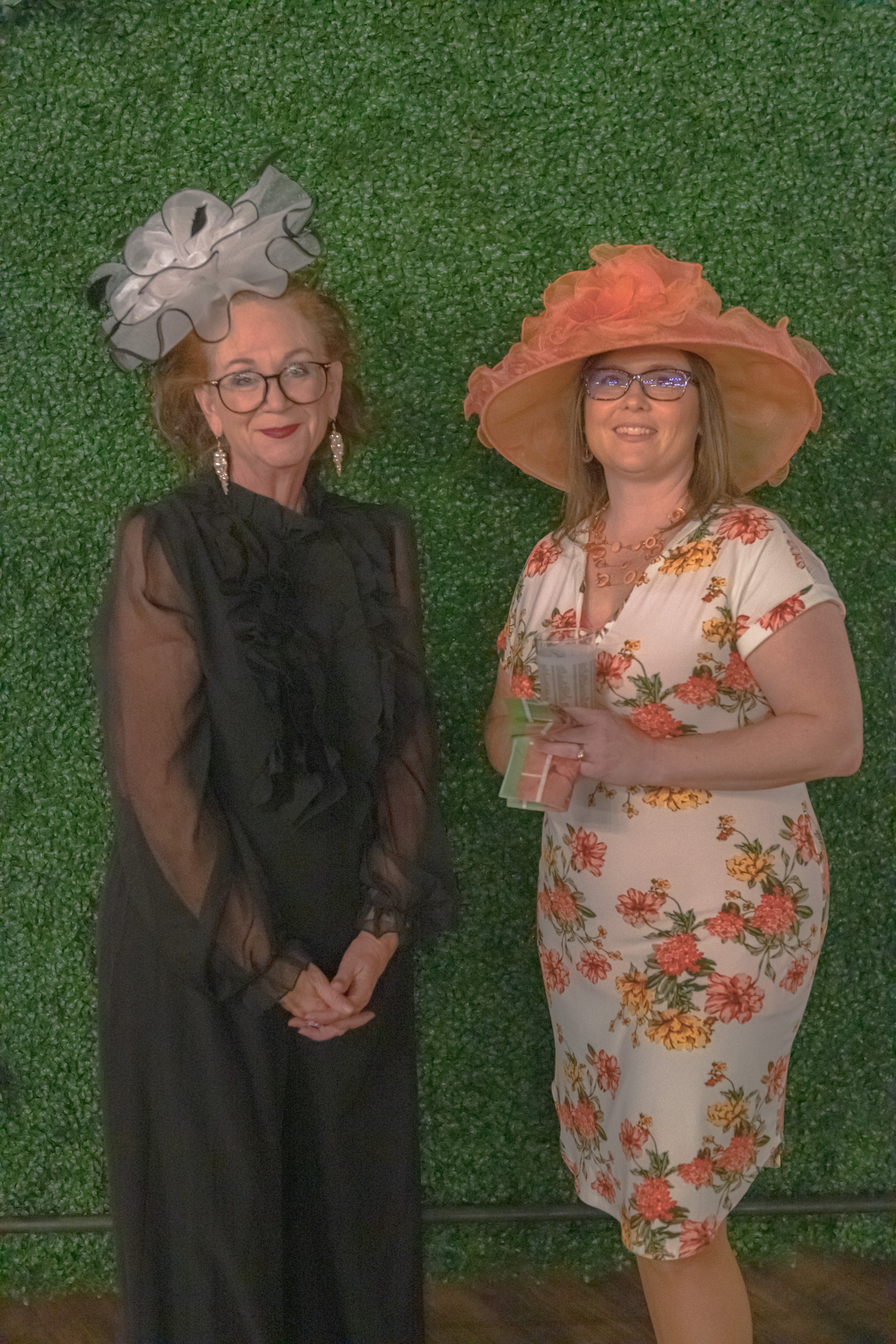 Two people pose against a green foliage backdrop, one in black with a silver headpiece and one in a floral dress and orange hat.