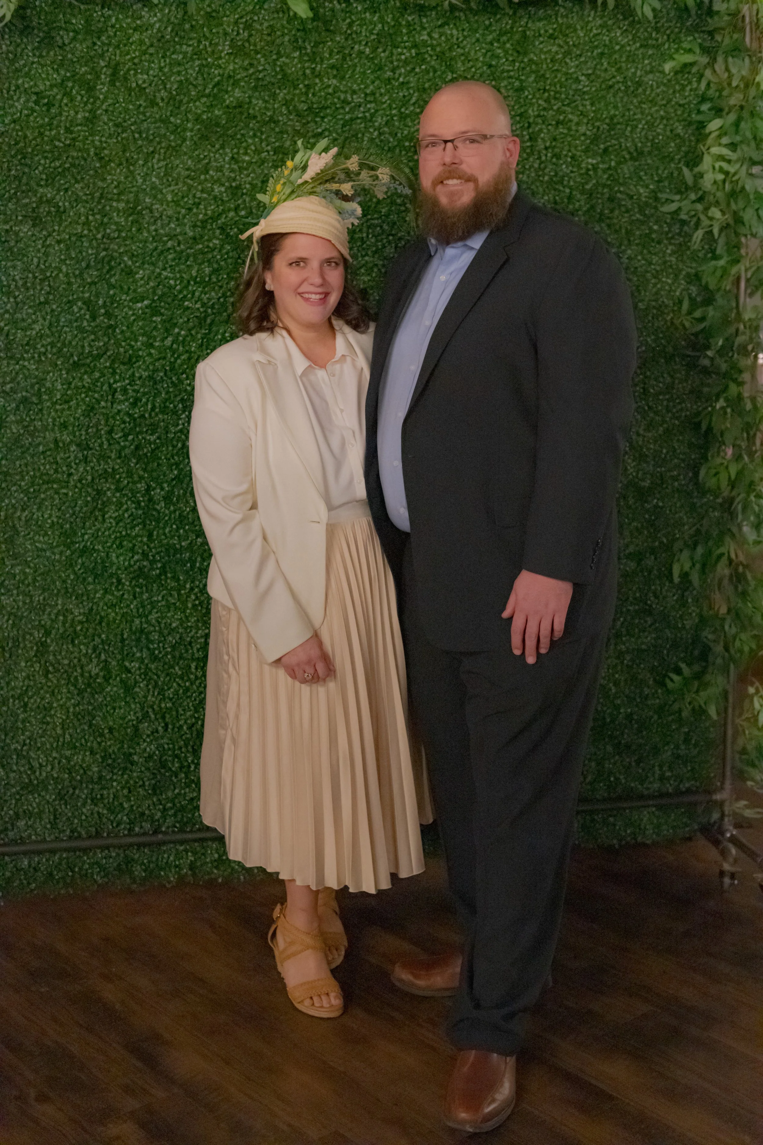 Full-length portrait of two people against a green foliage backdrop, one in a cream jacket and pleated skirt and one in a dark suit.